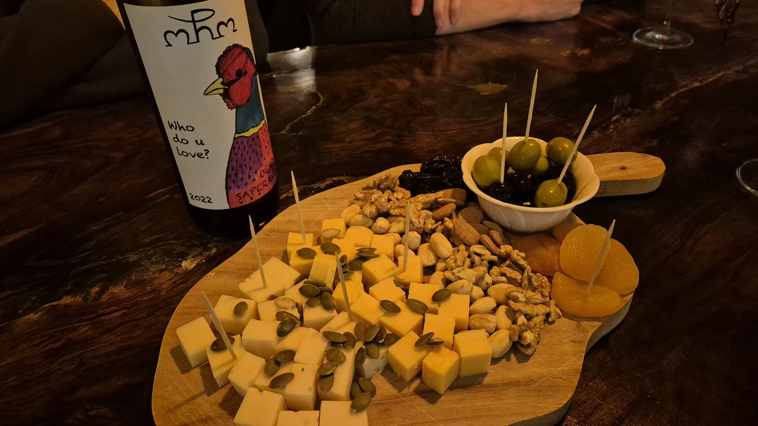 Wine Vibe Kutaisi a wooden board with several cubes of cheese, pumpkin seeds, a selection of nuts, olives and dried fruits. There is a bottle of natural red wine