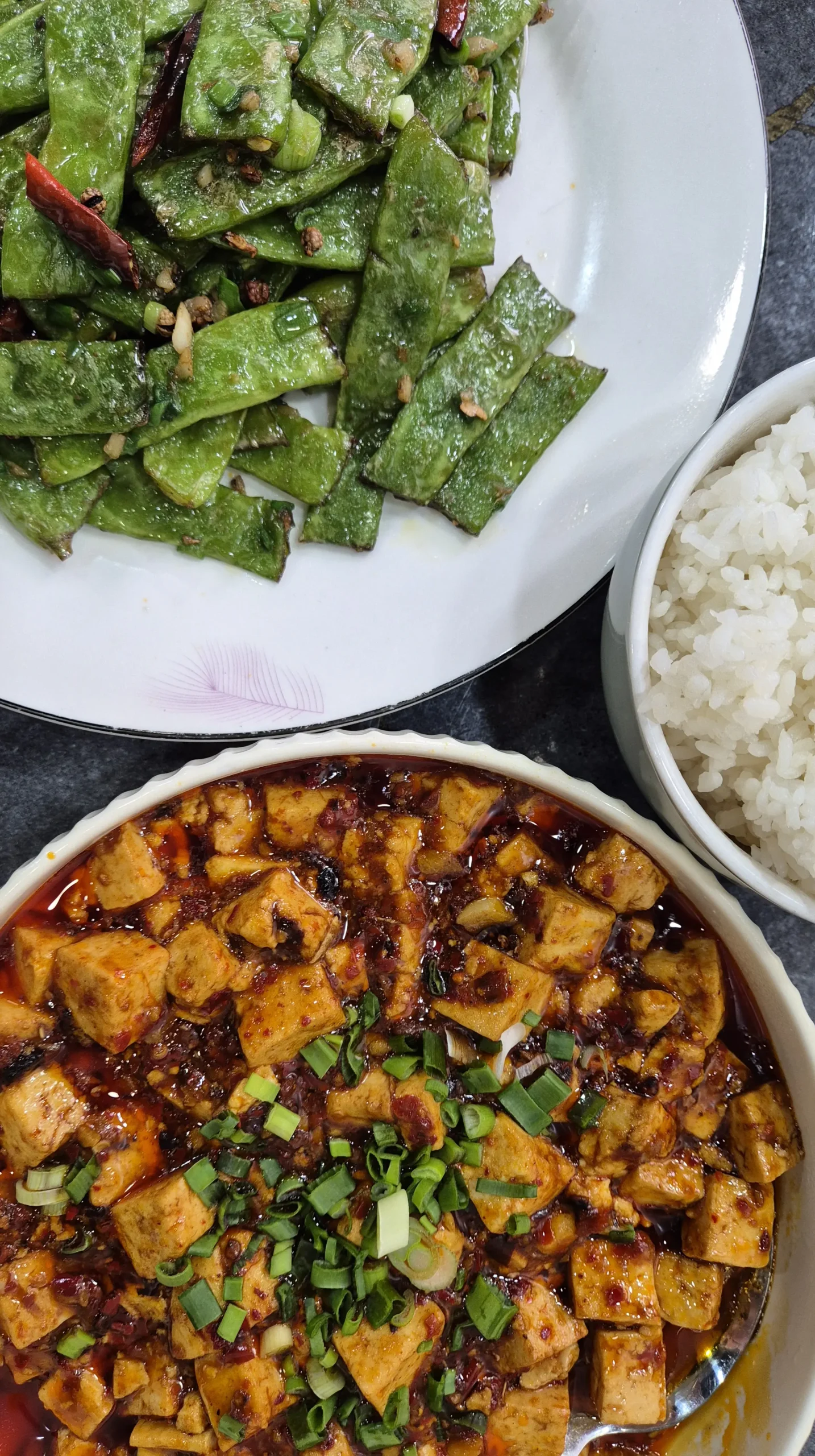 mapo tofu and spicy green beans three bowls sit next to each other on a table. Once has white rice, one has dry-fried, spicy green beans, and one has a bowl of spicy and saucy mapo tofu with spring onions on top