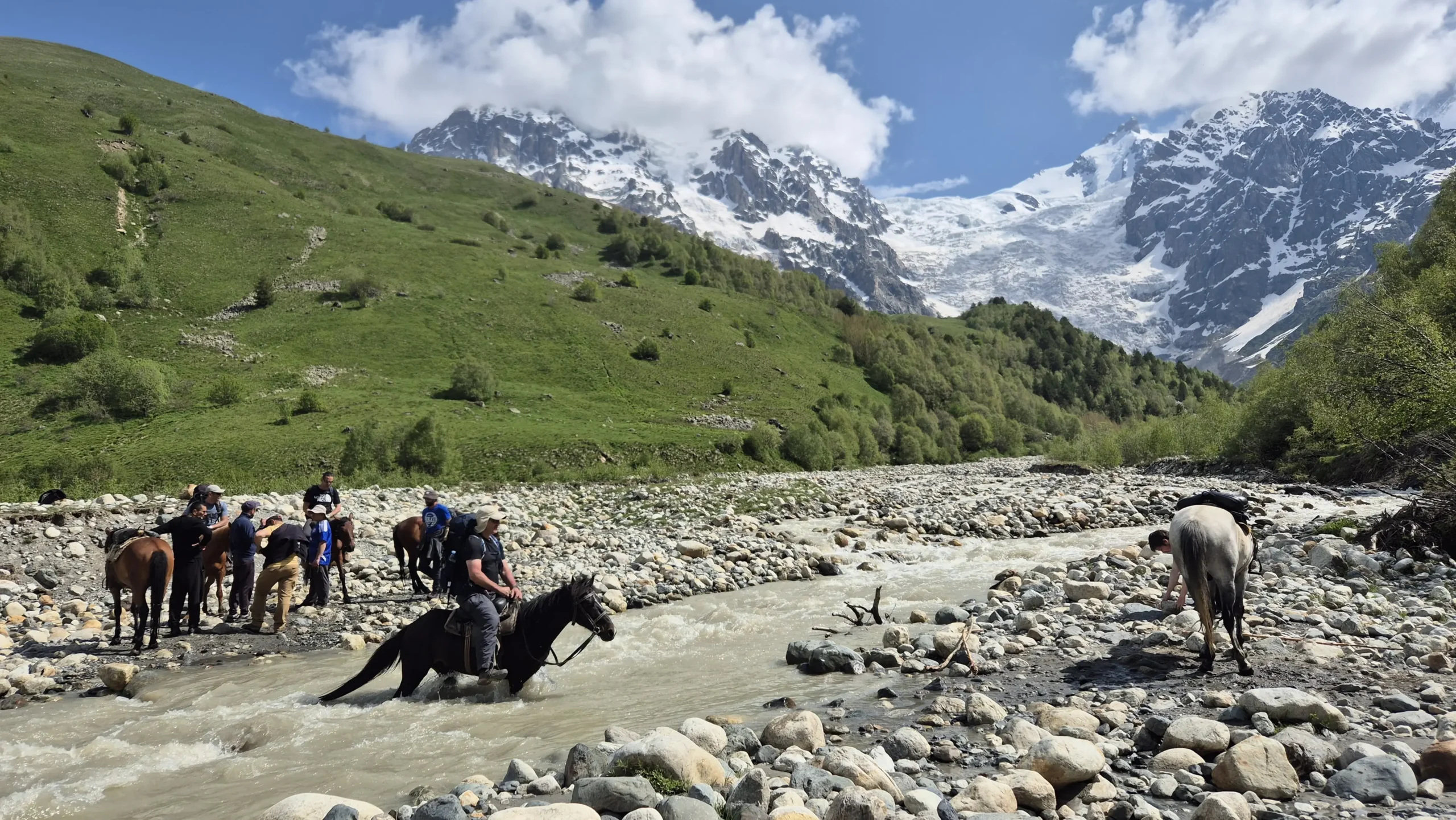 The infamous river crossing the infamous river crossing on leg 3 of the Mestia to Ushguli trek. A horse is carrying a man across a waist-deep river with mountains and a glacier in the background