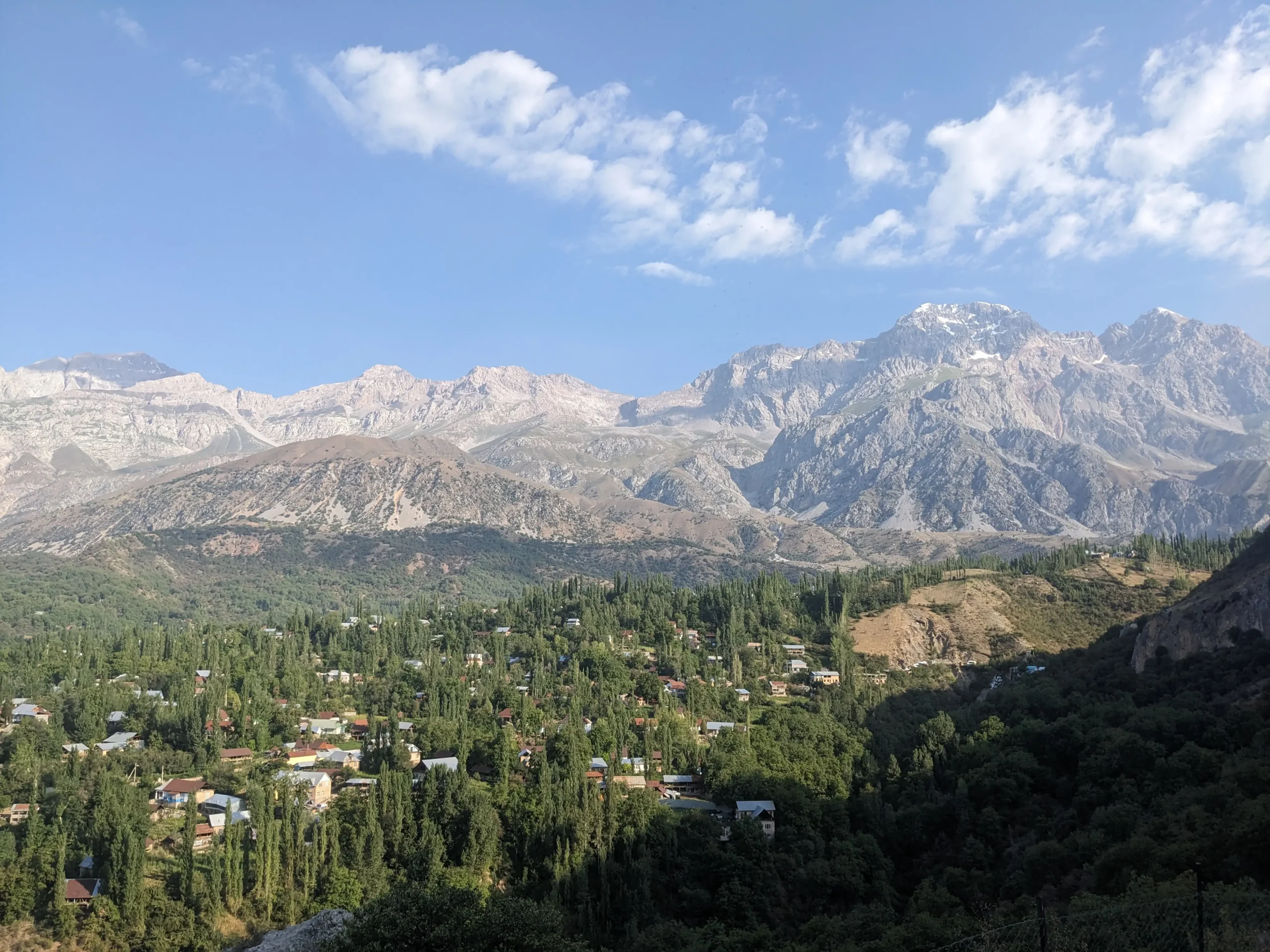 a view over a valley in Arslanbob in western Kyrgyzstan shows tall green trees with houses and a backdrop of huge grey mountains