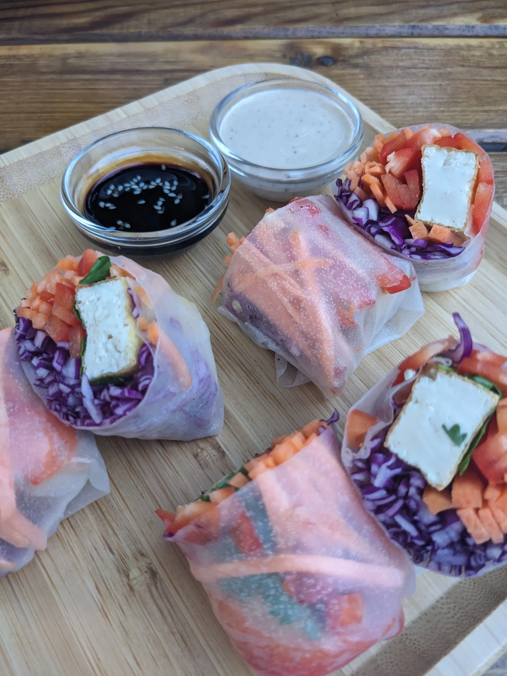 Summer rolls summer rolls stuffed with red cabbage, carrot, red pepper and tofu. Served on a wooden board alongside two dipping sauces - a cashew cream and a soy sauce