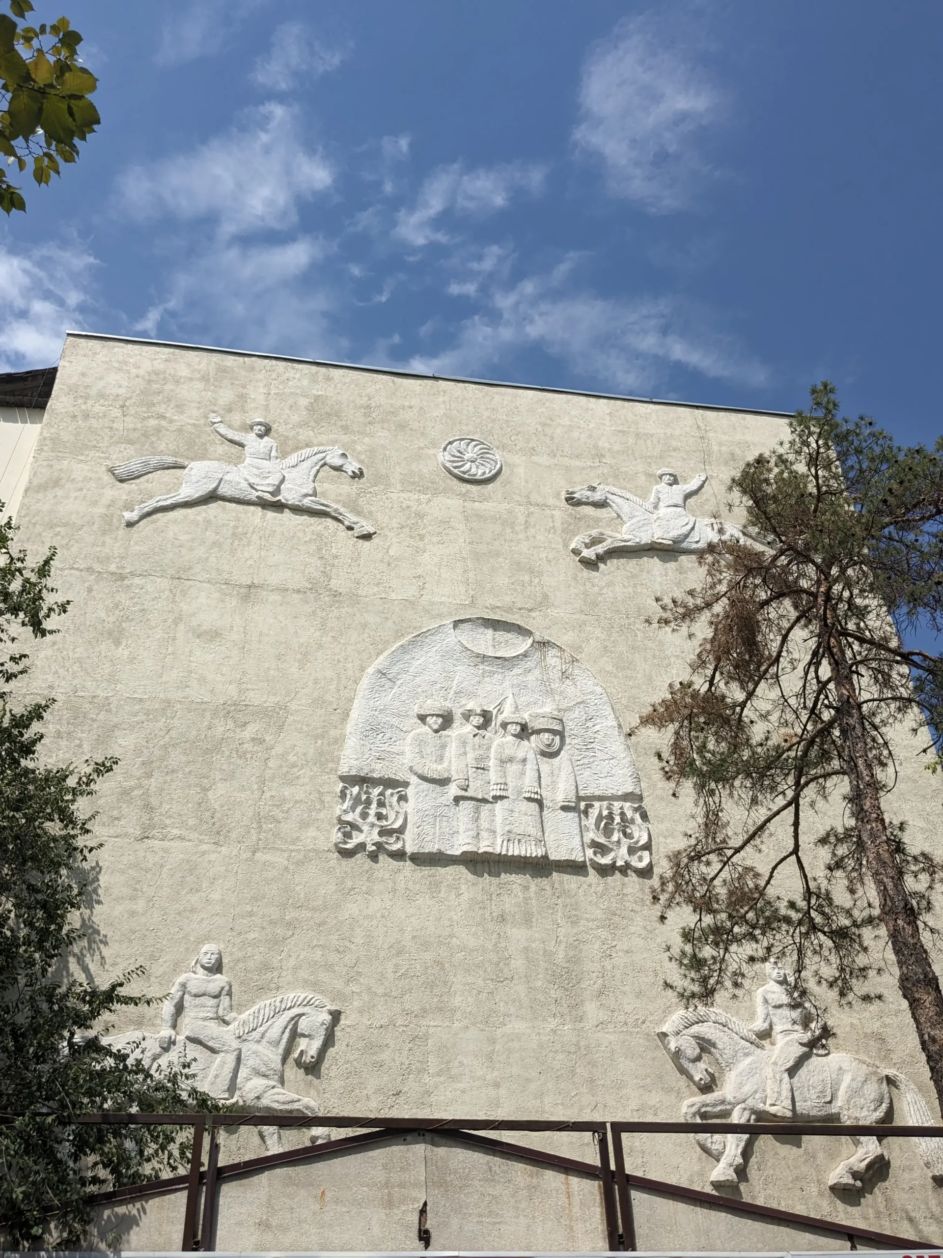 Yurt Bas Relief a white Soviet-style bas relief on the side of a building depicts local Kyrgyz people in local dress standing by a yurt. The sun is prominent and there are men riding horses. Bishkek, Kyrgyzstan