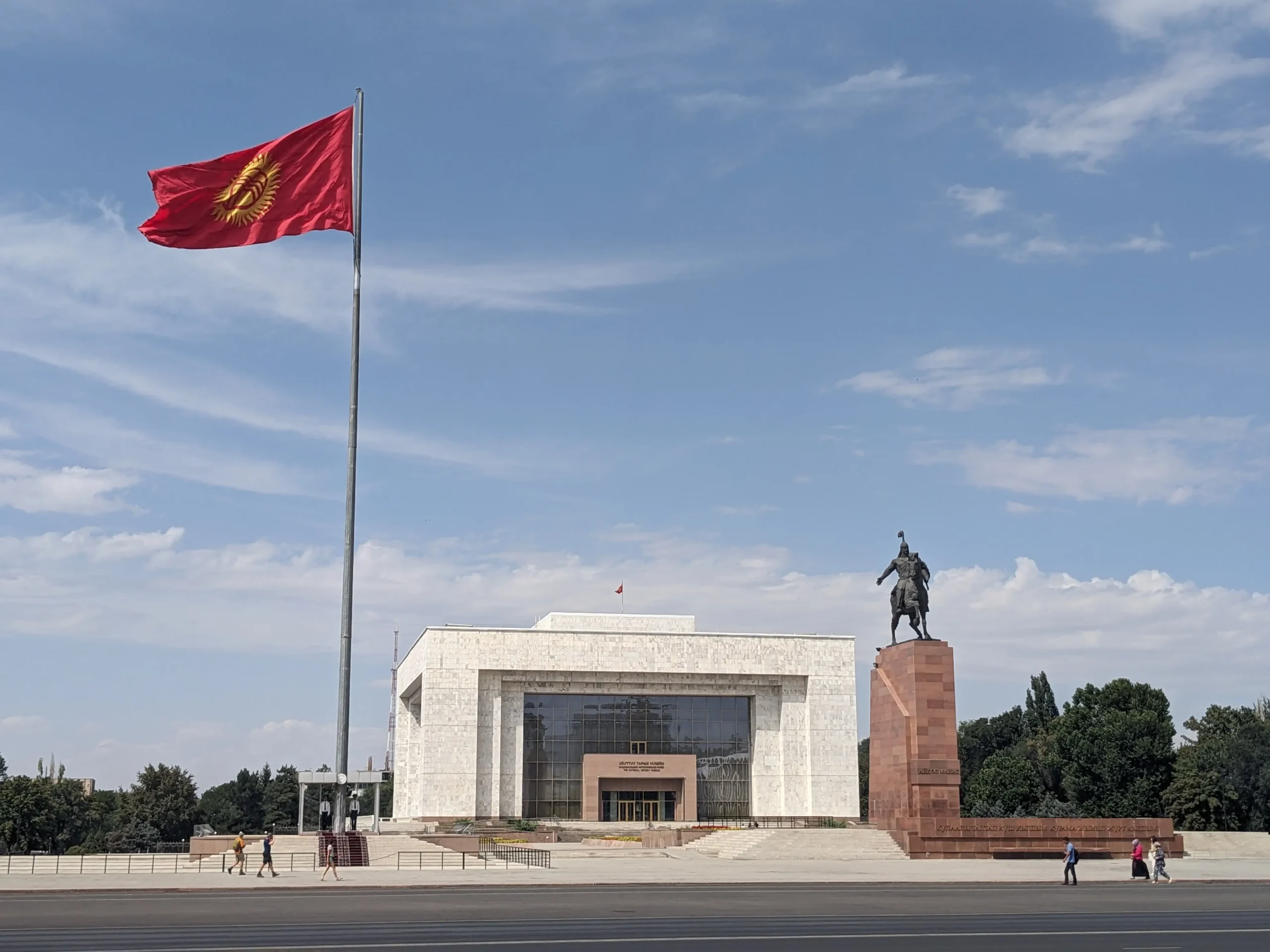 a photo of the history museum in Bishkek which is housed in a large white marble rectangular building in Soviet modernist style. There is a large Kyrgyz flag pole and a statue.