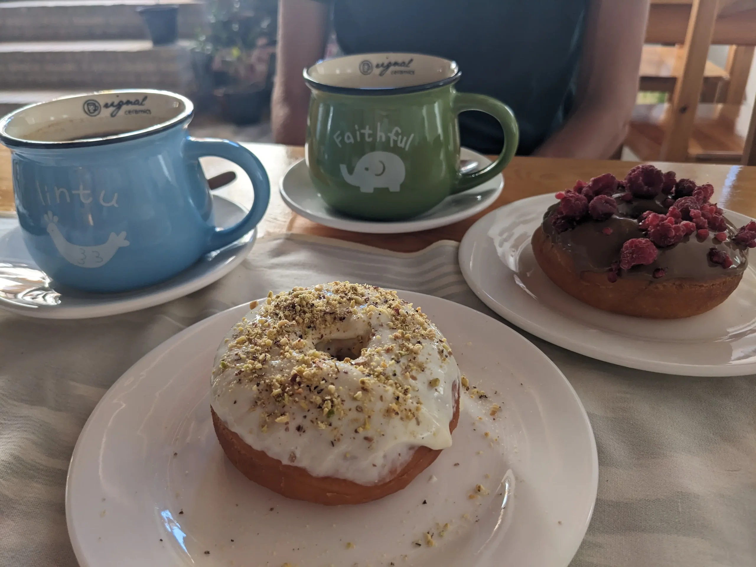 Donuts and Coffee two cute, fat mugs in green and blue has a bird and an elephant on. alongside the mugs are two plates of donuts - one covered in chocolate and raspberries, the other with white chocolate and crushed nuts in Bukhara, Uzbekistan - Donuts and Coffee cafe