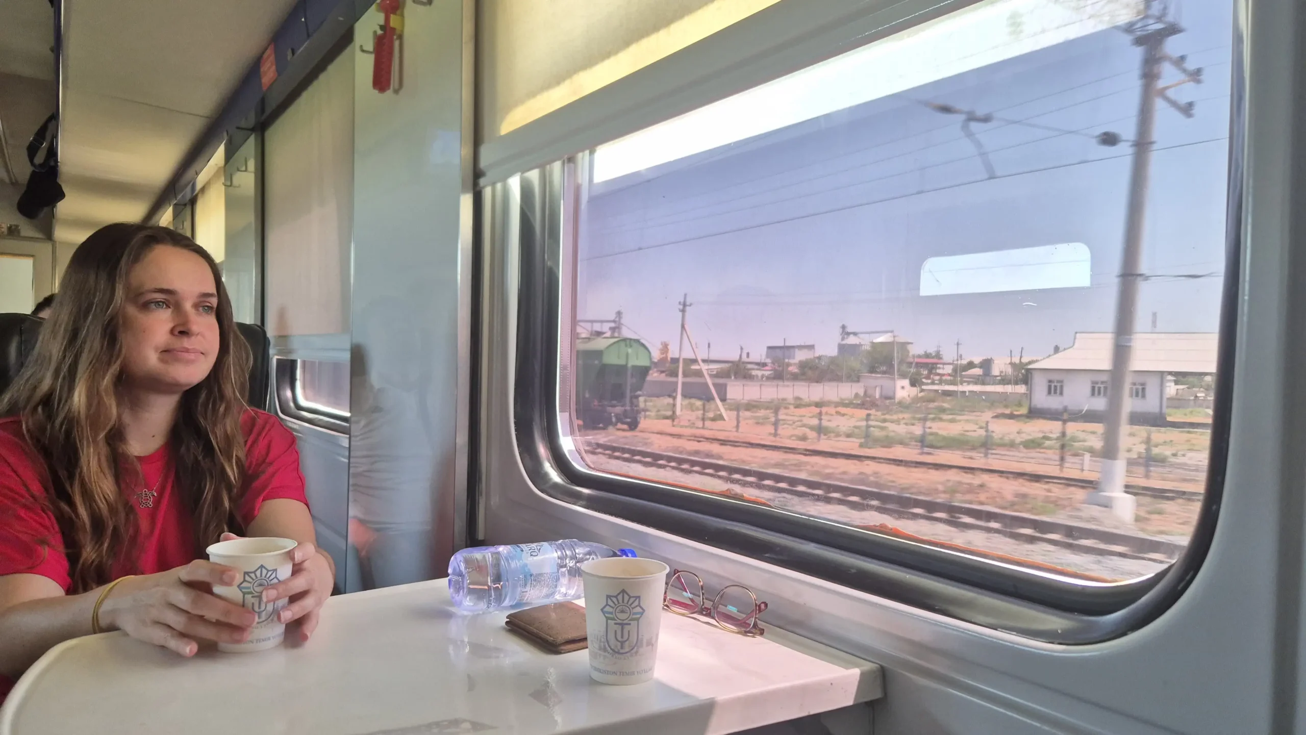 a woman is sitting on a train holding a cup of tea and looking out the window. Andijan to Osh