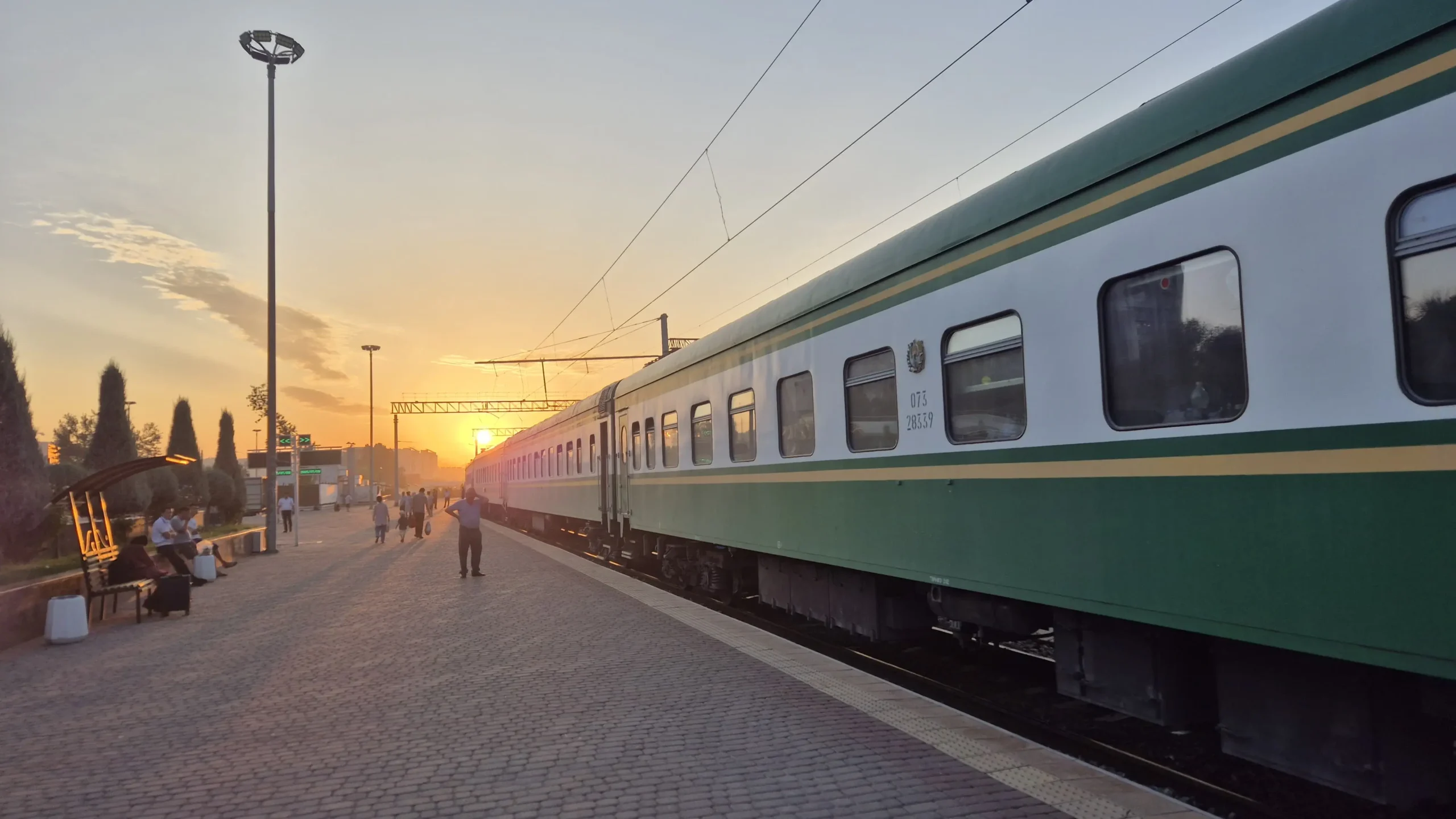 Uzbek sleeper train the sun is rising at a train station where a white and green sleeper train is waiting at the platform