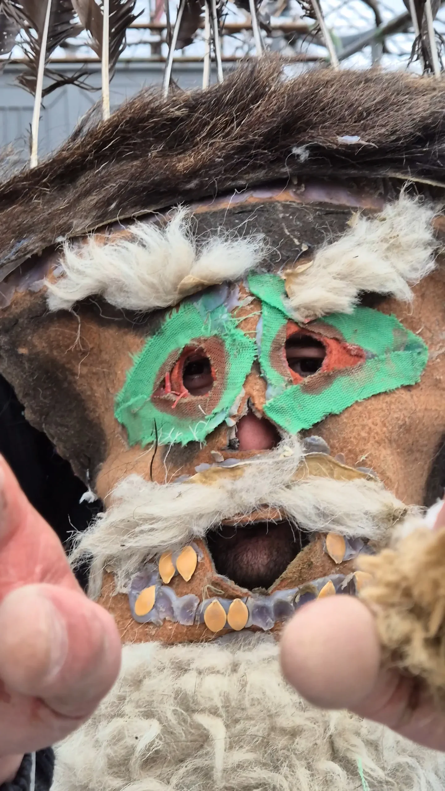 Berika! a man dressed up as a berika during Berikaoba Pagan festival held in Didi Chailuri, Georgia. he is wearing a brown mask with green and orange material around the eyes, white material for the eyebrows and moustache, and rows of pumpkin seeds for teeth