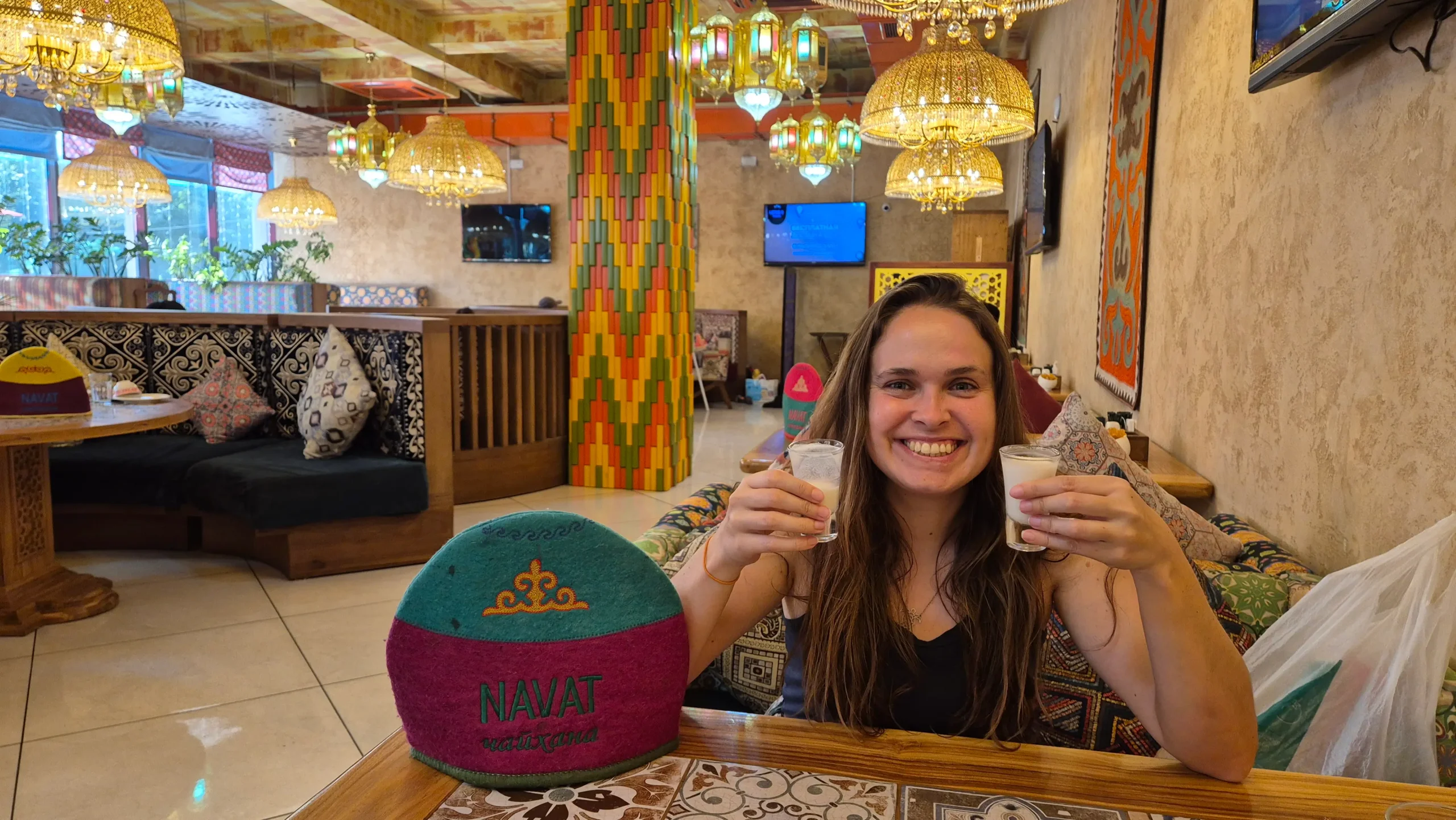 a woman holds up two small glasses of chalap (fermented milk) in a restaurant decorated with large, extravagant lampshades - Navat in Osh, Kyrgyzstan