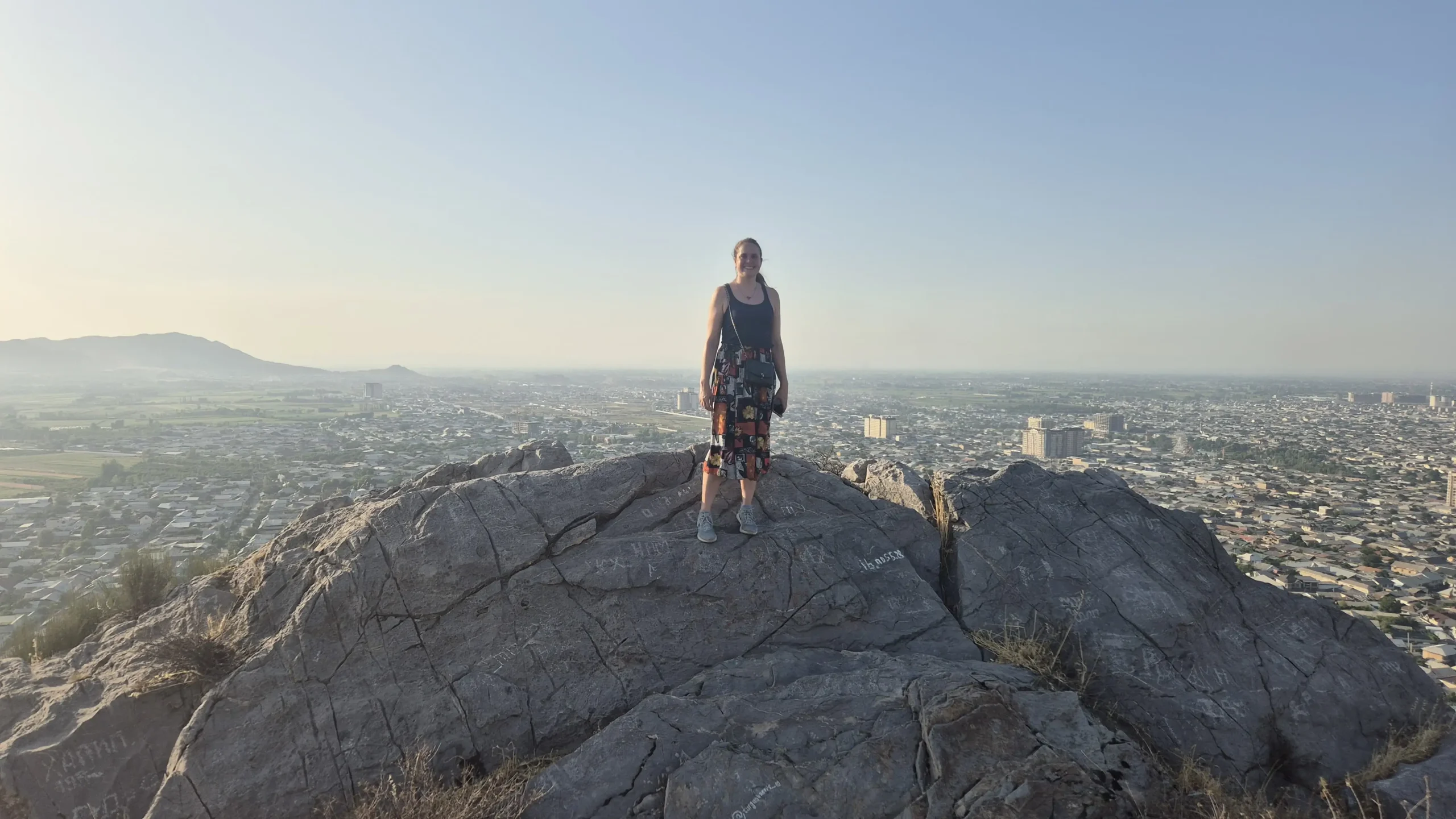a woman stands on a large rock at sunset with a view over Osh city in the background, Kyrgyzstan