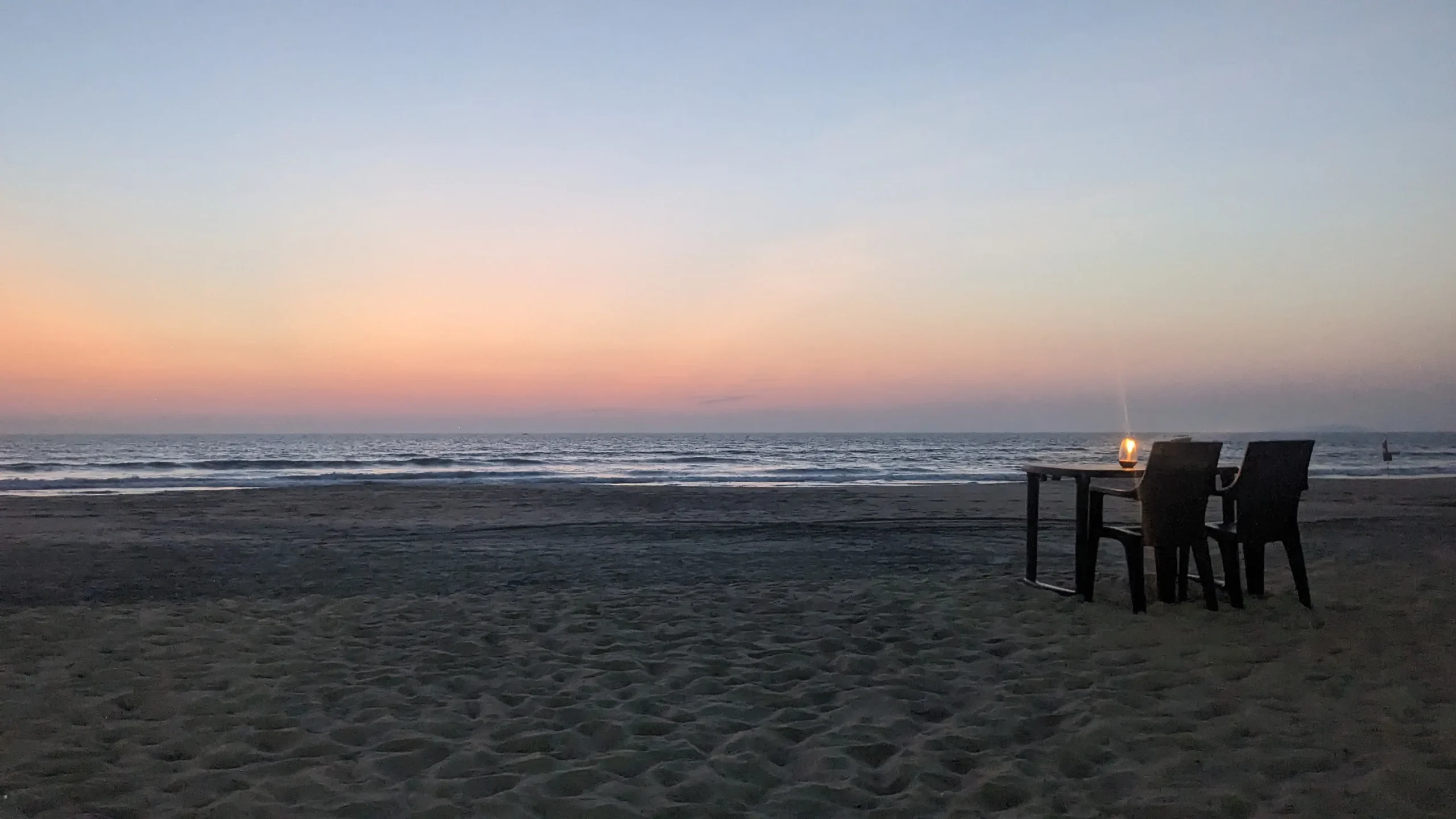 Two seats, and a table with a lit candle sit on the sand looking out over pastel coloured skies at sunset on Majorda Beach, North or South Goa