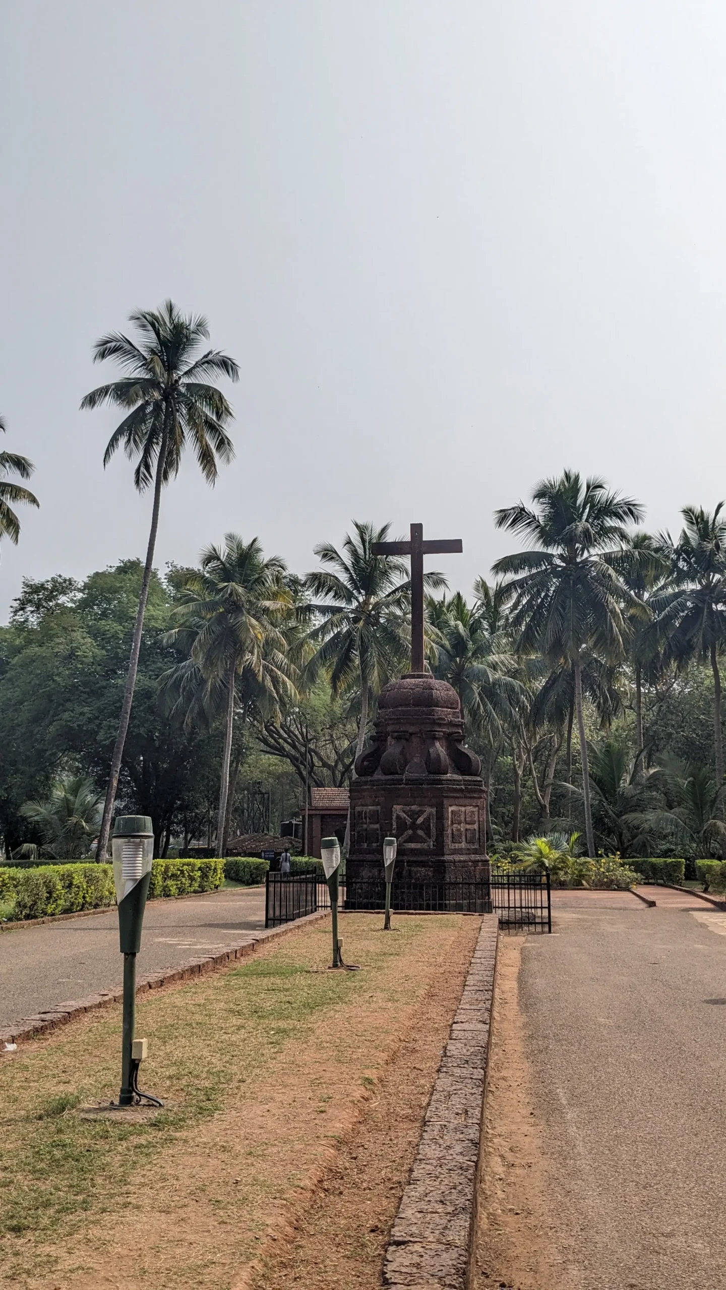 a religious cross with a short top on a brick pedestal surrounded by palm trees outside of Basilica do Bom Jesus in Old Goa, India
