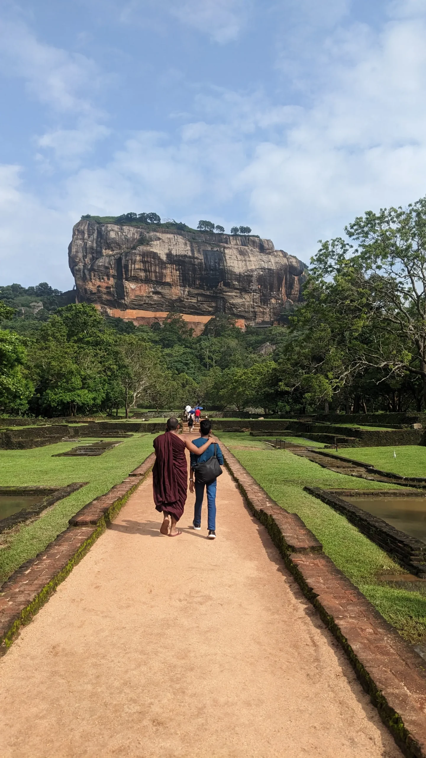 a paved walkway surrounded by maintained gardens leads towards the huge granite rock of Sigiriya Rock.