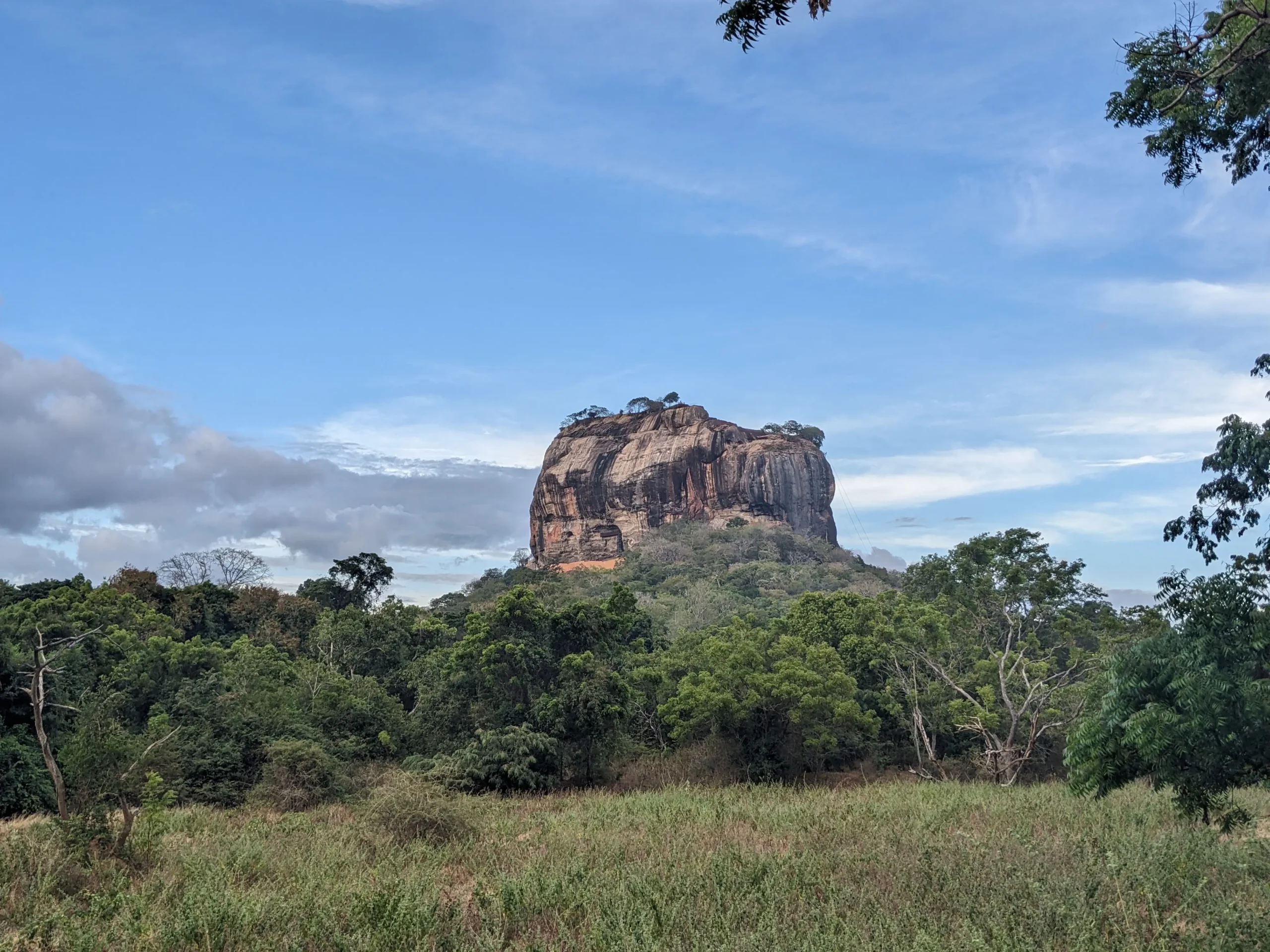 a huge 180m tall granite rock stands on the plains of Sigiriya in Sri Lanka surrounded by trees. 