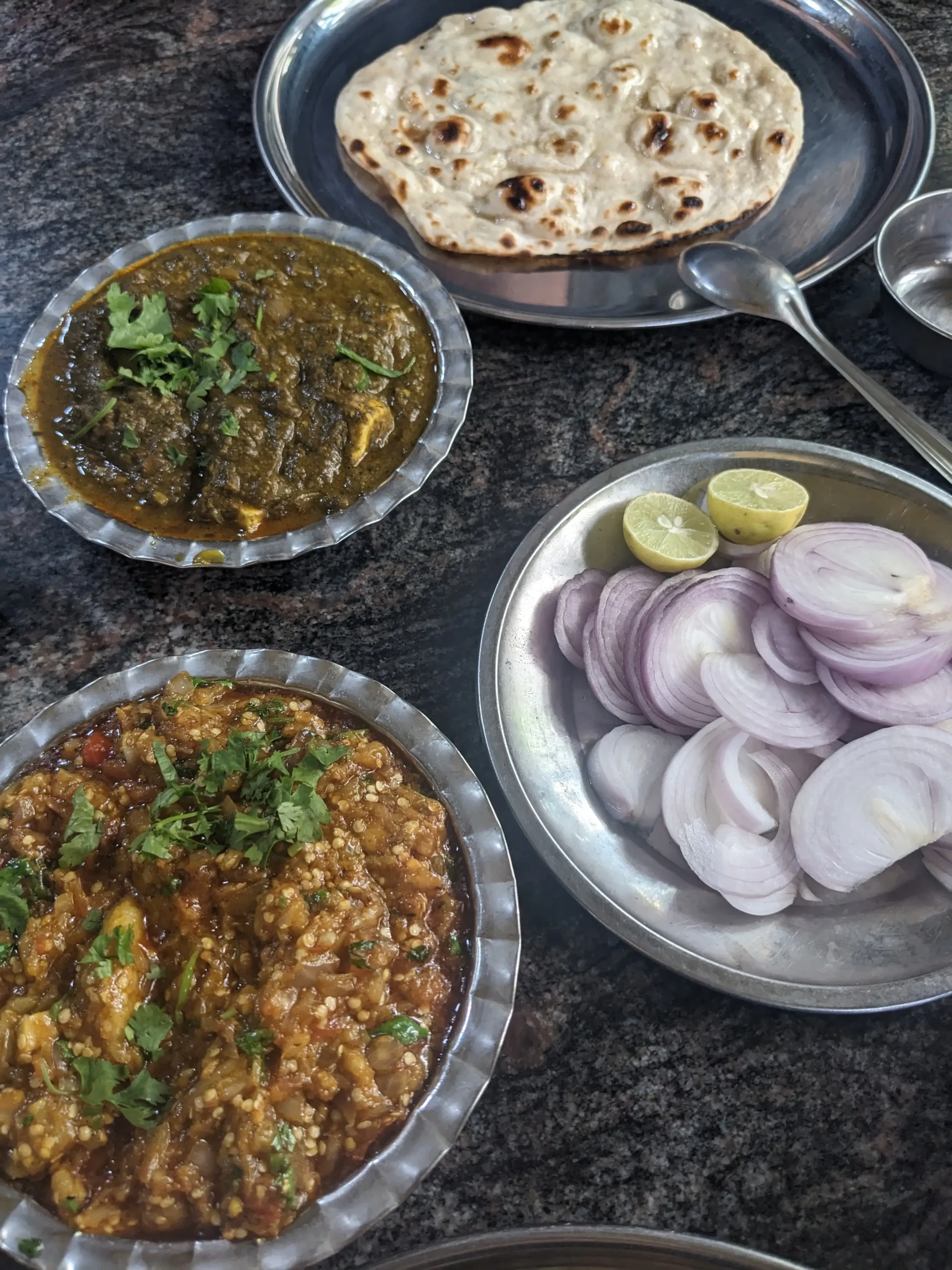 four metal bowls on a table. One contains a deep green curry (palak paneer), one has spiced and mashed aubergine (baingan bharta), one has sliced red onions and one has bread.