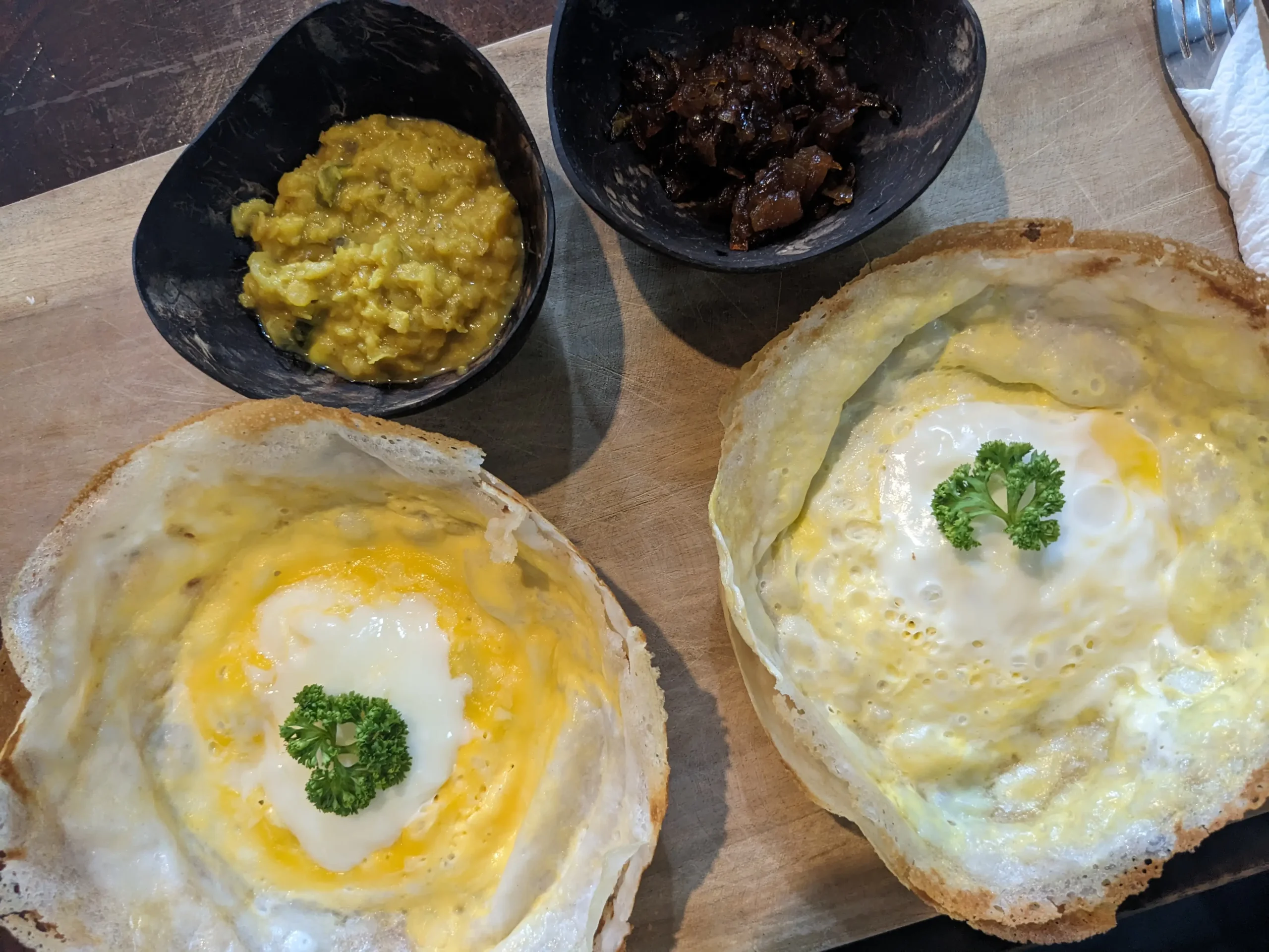 Egg hoppers, dal, & onion chutney two Sri Lankan egg hoppers (thin savoury pancake with a whole egg cooked at the bottom) sit on a wooden board with a bowl of dal and onion chutney