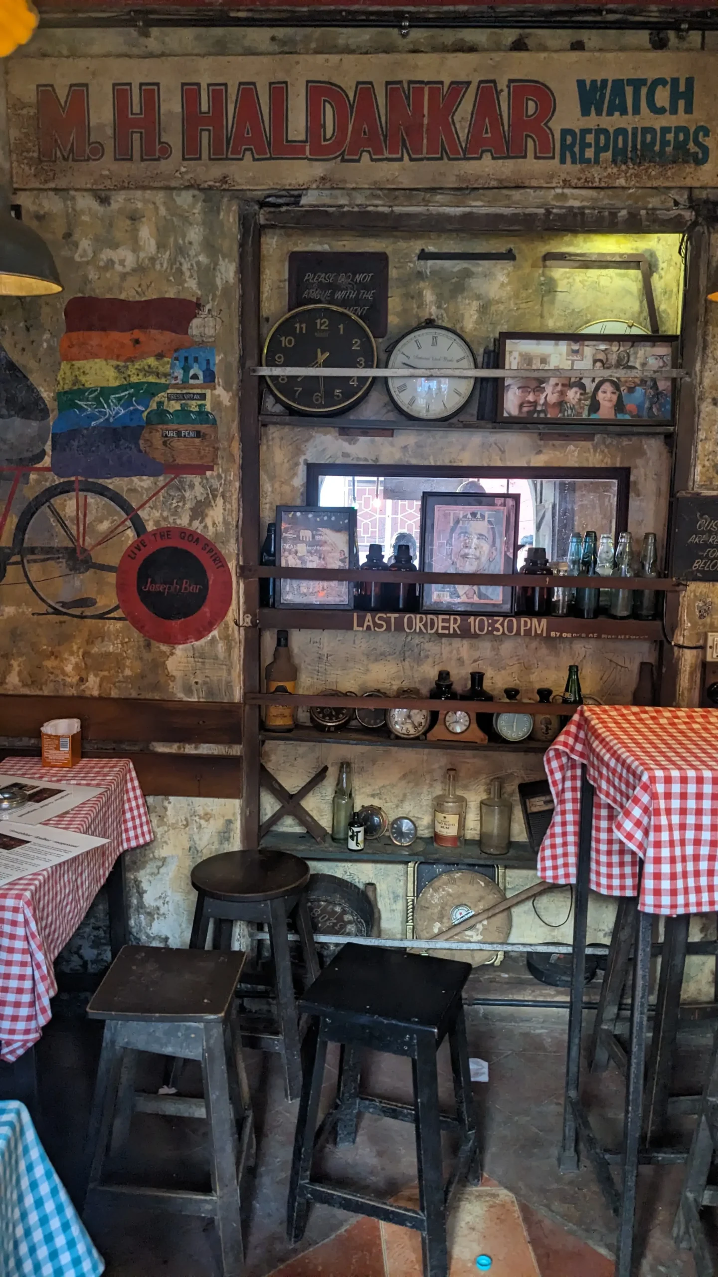Joseph's Bar interior the retro interior of a bar that used to be a watch repair shop has an old sign, old clocks, bottles, and photos on wooden shelves. There are wooden chairs and table with red and white chequered table cloth - Joseph Bar, Panaji, Goa, India