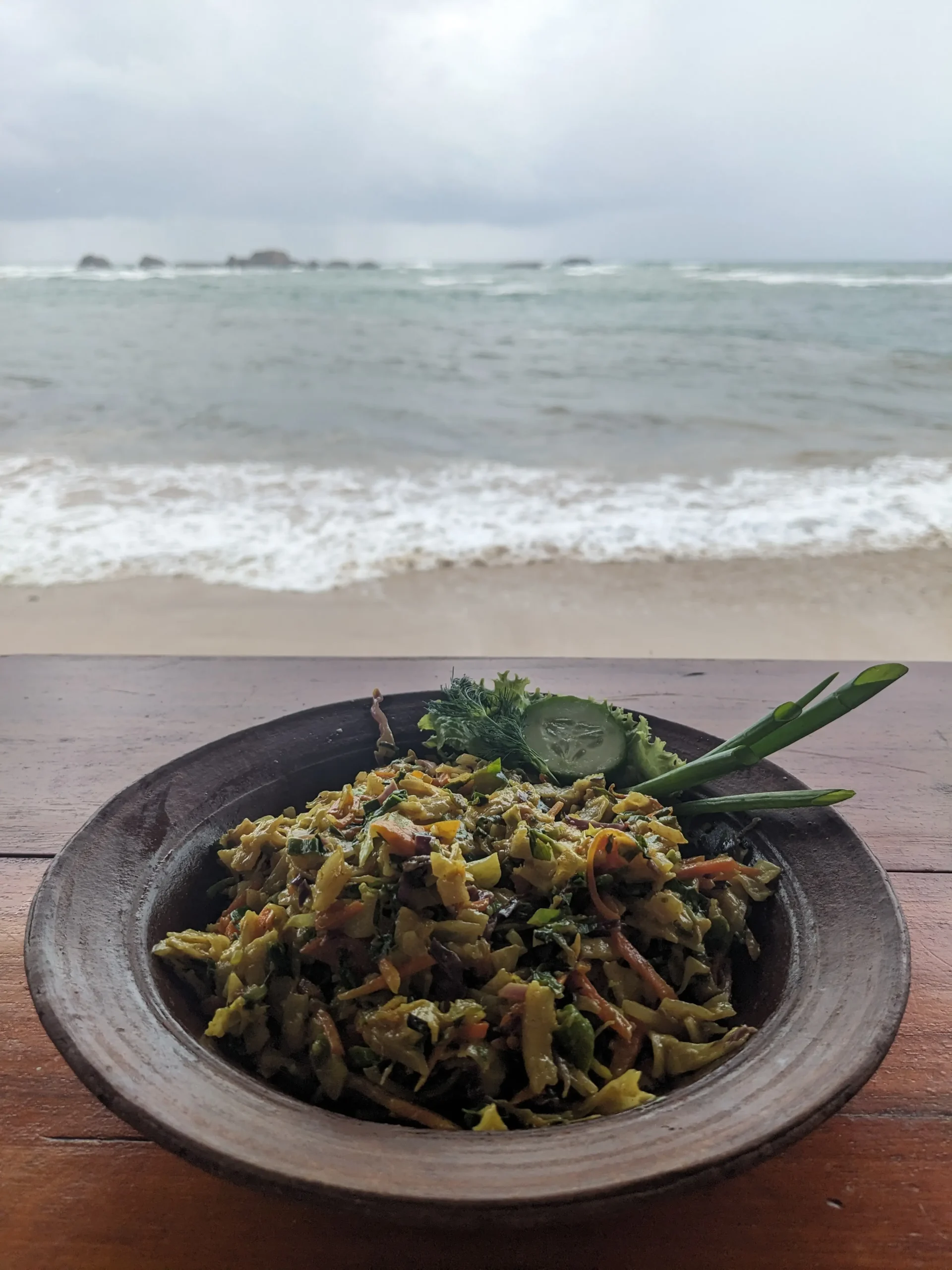 Kottu roti with a sea view a clay bowl of kottu roti (chewy flatbread chopped into small pieces and mixed with veg and curry sauce) sits on a wooden table overlooking the sea.