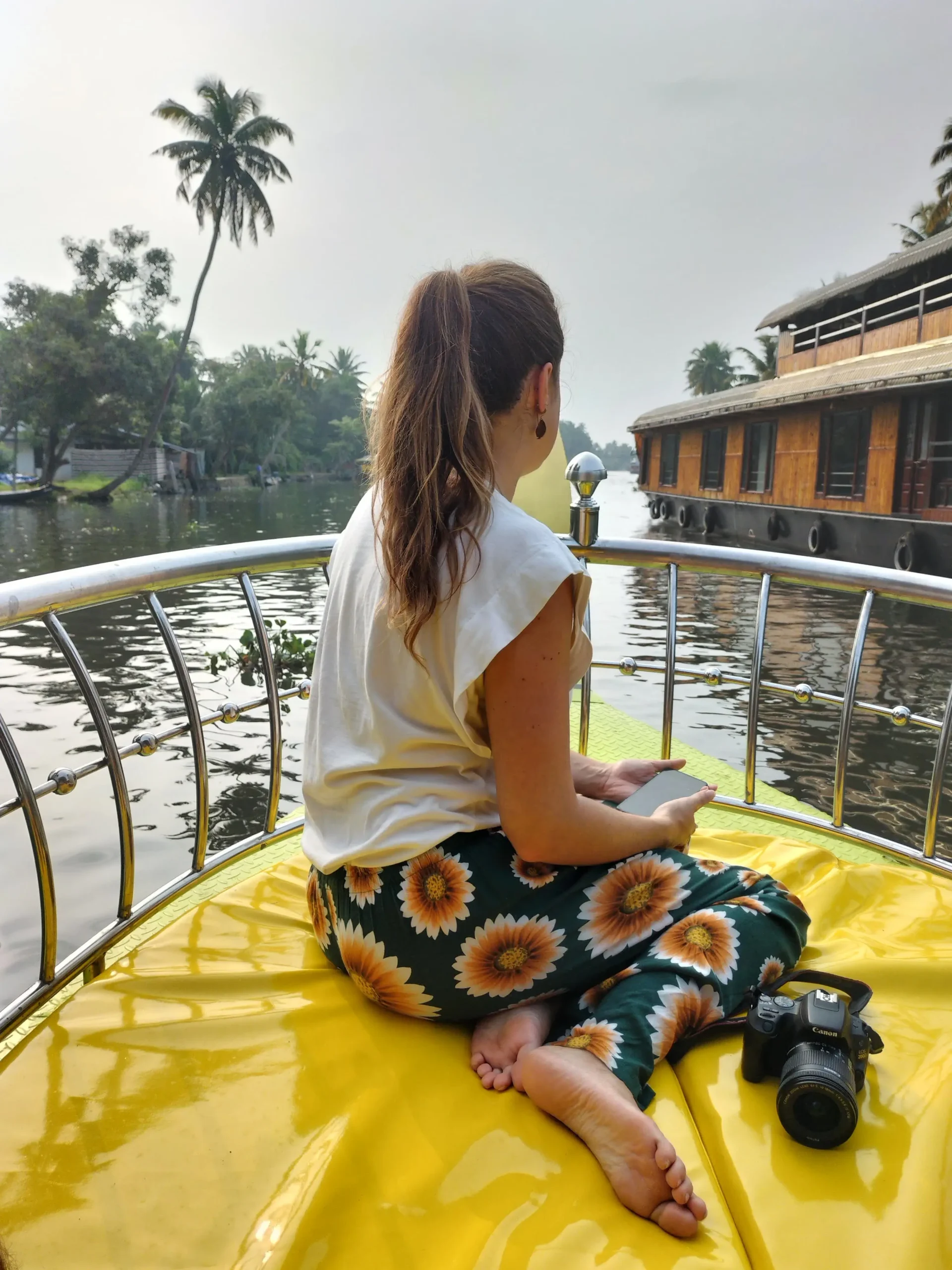 a woman is sat at the front of a boat  floating through the backwaters of Alleppey, Kerala, South India. The water is lined with palm trees
