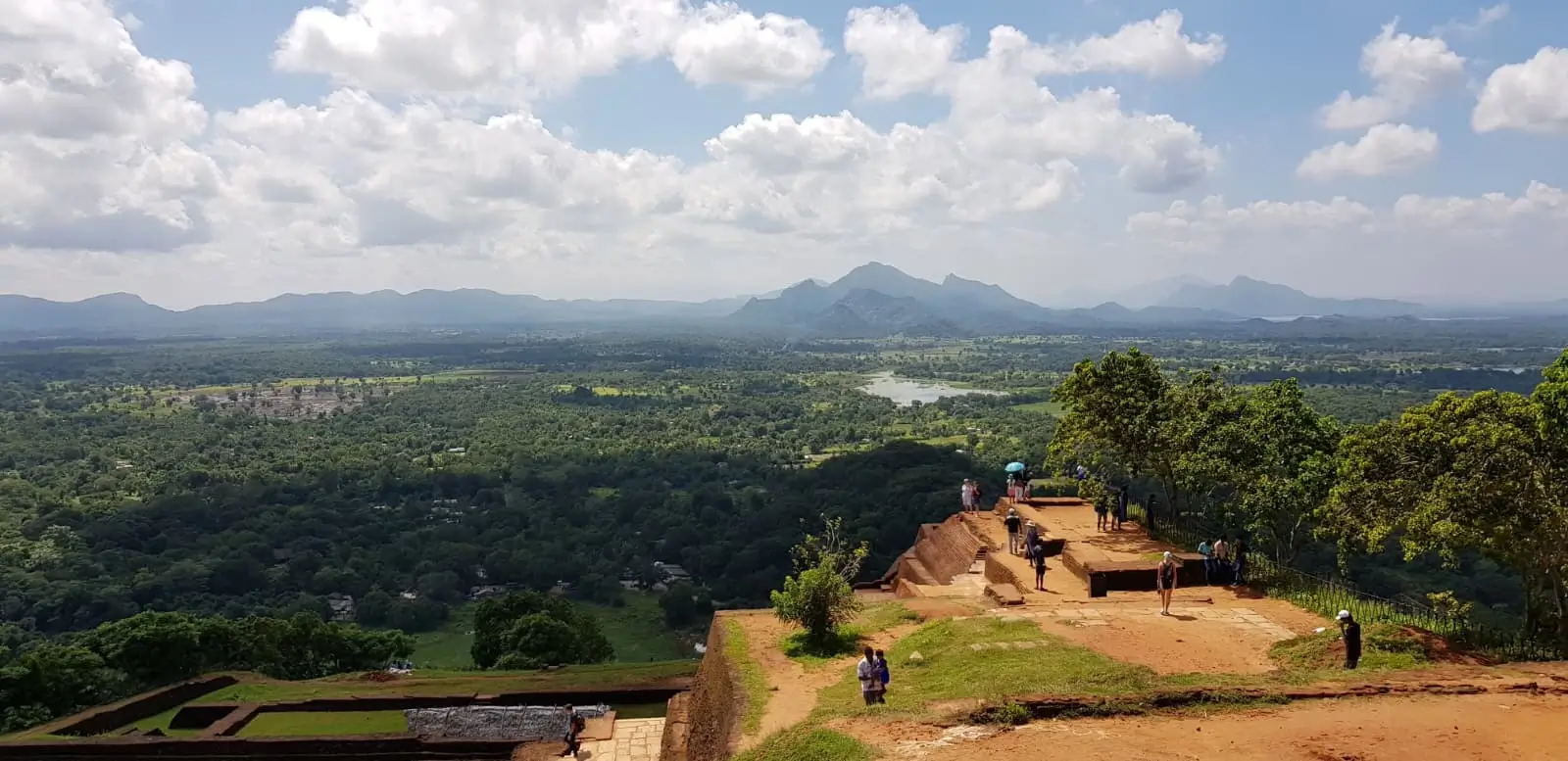 the view from the top of Sigiriya Rock over the jungle with a lake, and mountains in the background, and ruins of an ancient citadel in the foreground