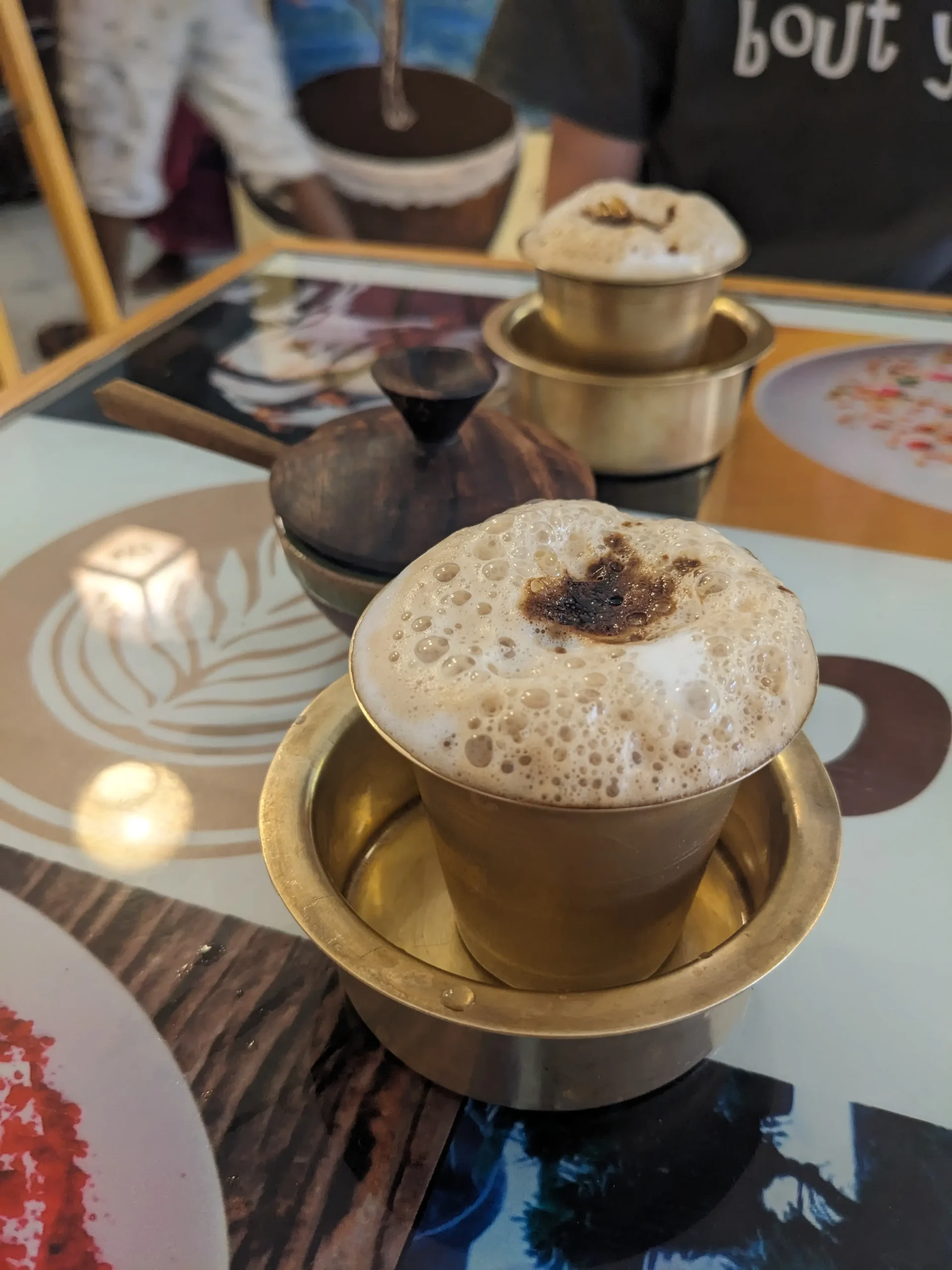 a gold metal cup full of frothy South indian filter coffee sits inside a wide gold metal bowl