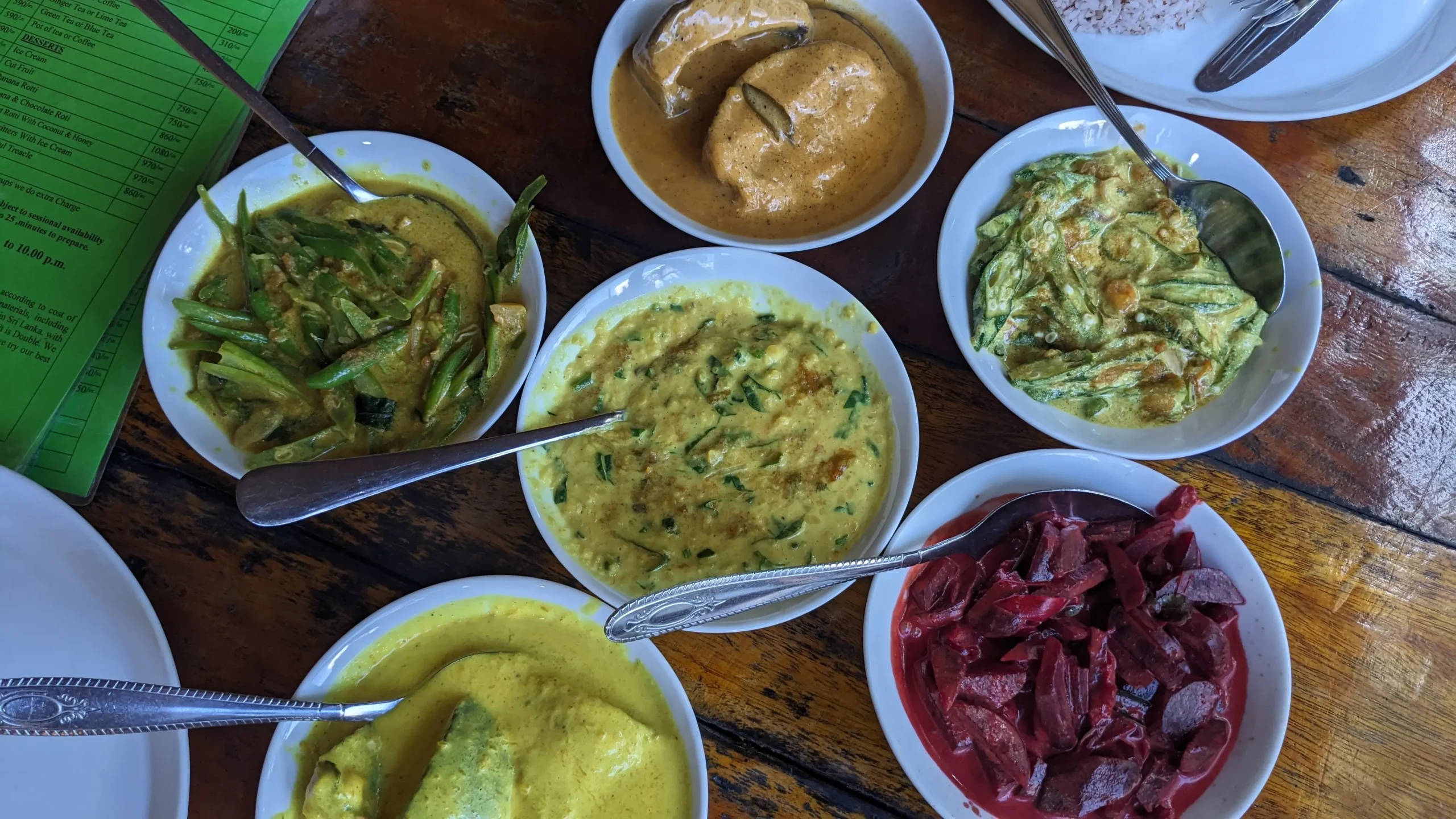 six bowls of Sri Lankan curries on a table - an orange mango curry, a red beetroot curry, a yellow okra curry, a dal, a yellow green bean curry, a yellow pumpkin curry