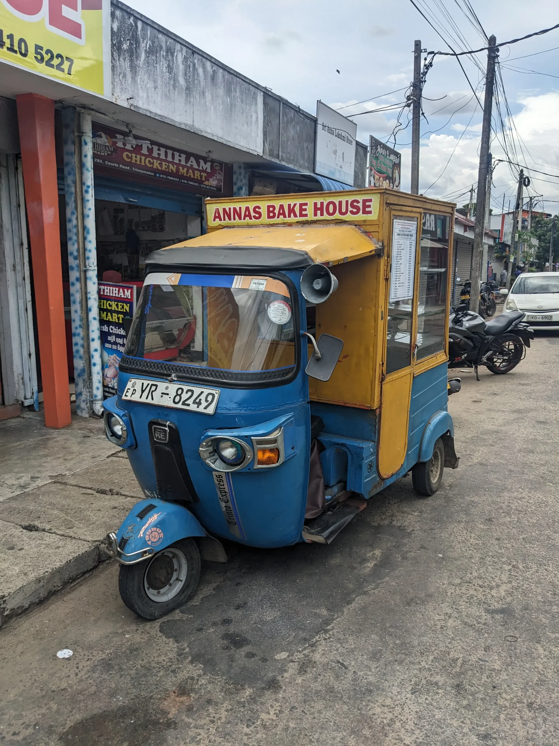 a blue and yellow tuktuk has the back seats blocked off and turned into shelving with a glass window to house baked goods such as veg curry buns - Sri Lankan singing bread van