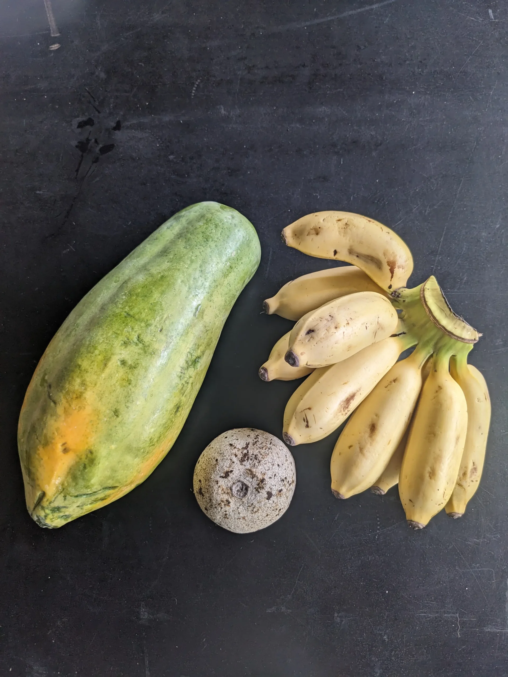 a green papaya sits on a black table next to a bunch of small Sri Lankan bananas and a wood apple
