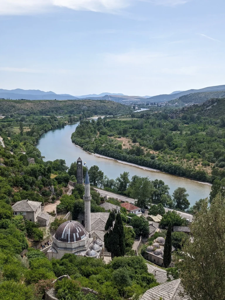 a historic Ottoman style mosque with several domes sits beside a crumbling hammam and other stone buildings surrounded by green trees, while the blue water of Neretva river flows past. Počitelj, Bosnia and Herzegovina