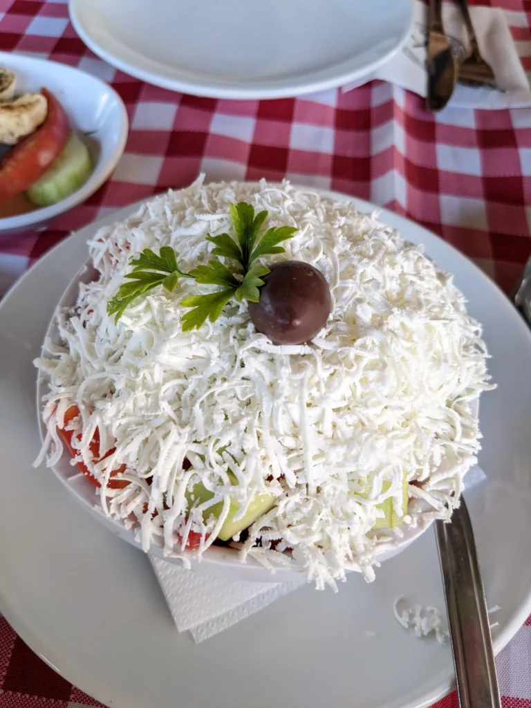 a bowl of shopska salad is made up of tomato and cucumber heavily topped with white grated cheese, a black olive and sprig of parsley in Bosnia and Herzegovina