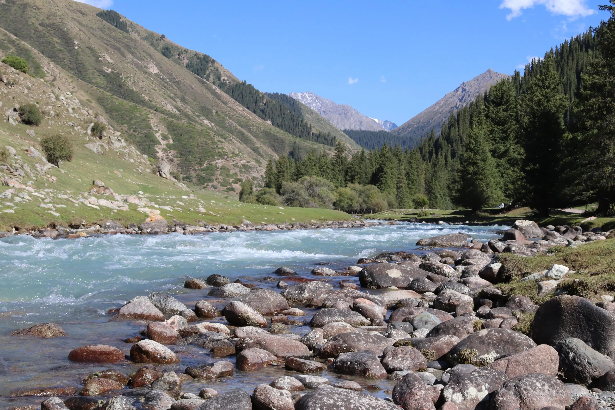 a view up the mountain river in Jeti Oguz Gorge, Kyrgyzstan. There are large pebbles in the foreground, blue river water, green trees and mountains in the background