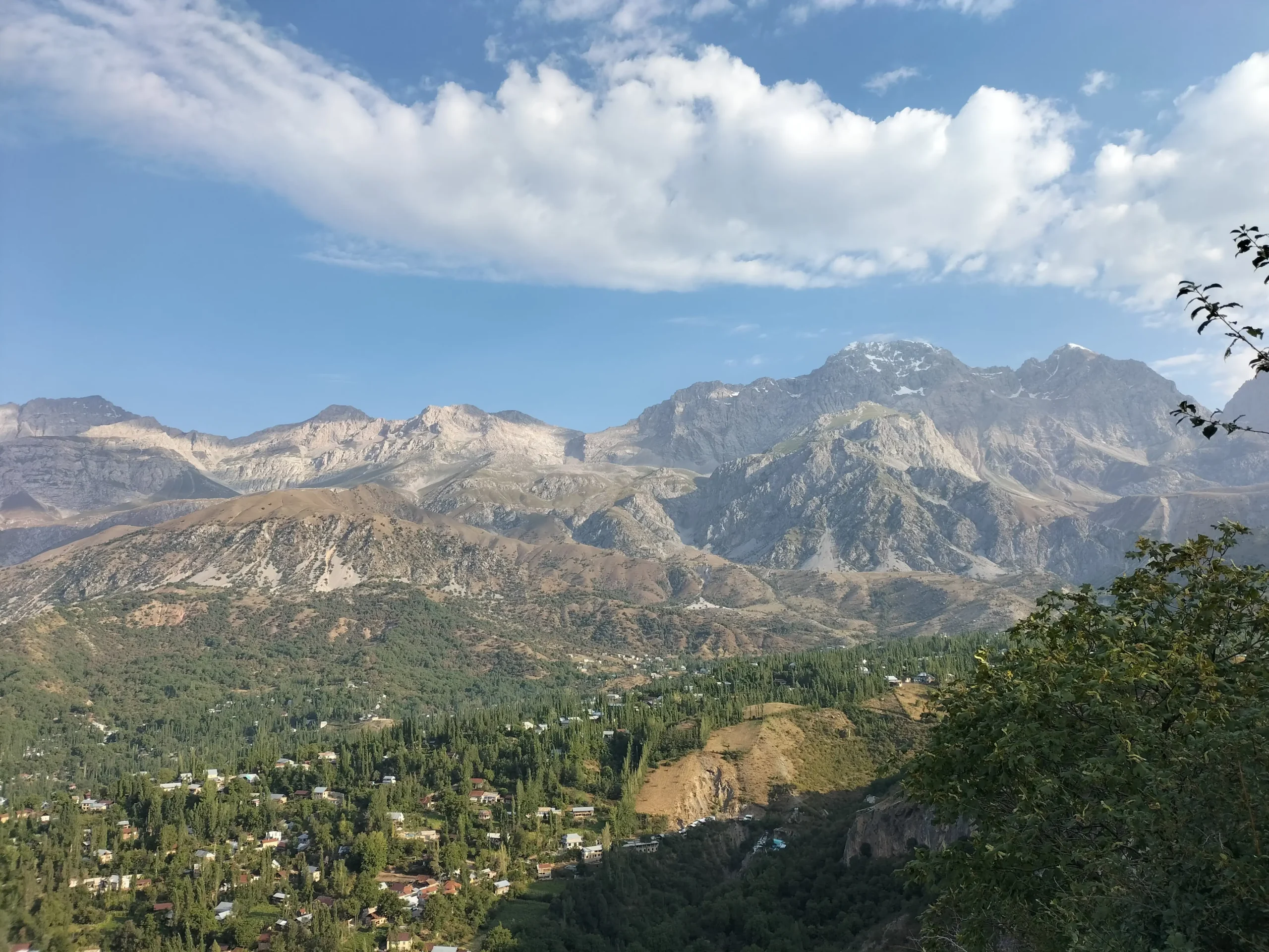 wild, rugged mountains of Arslanbob, Kyrgyzstan have tall trees in the lower ground in front with housing interspersed