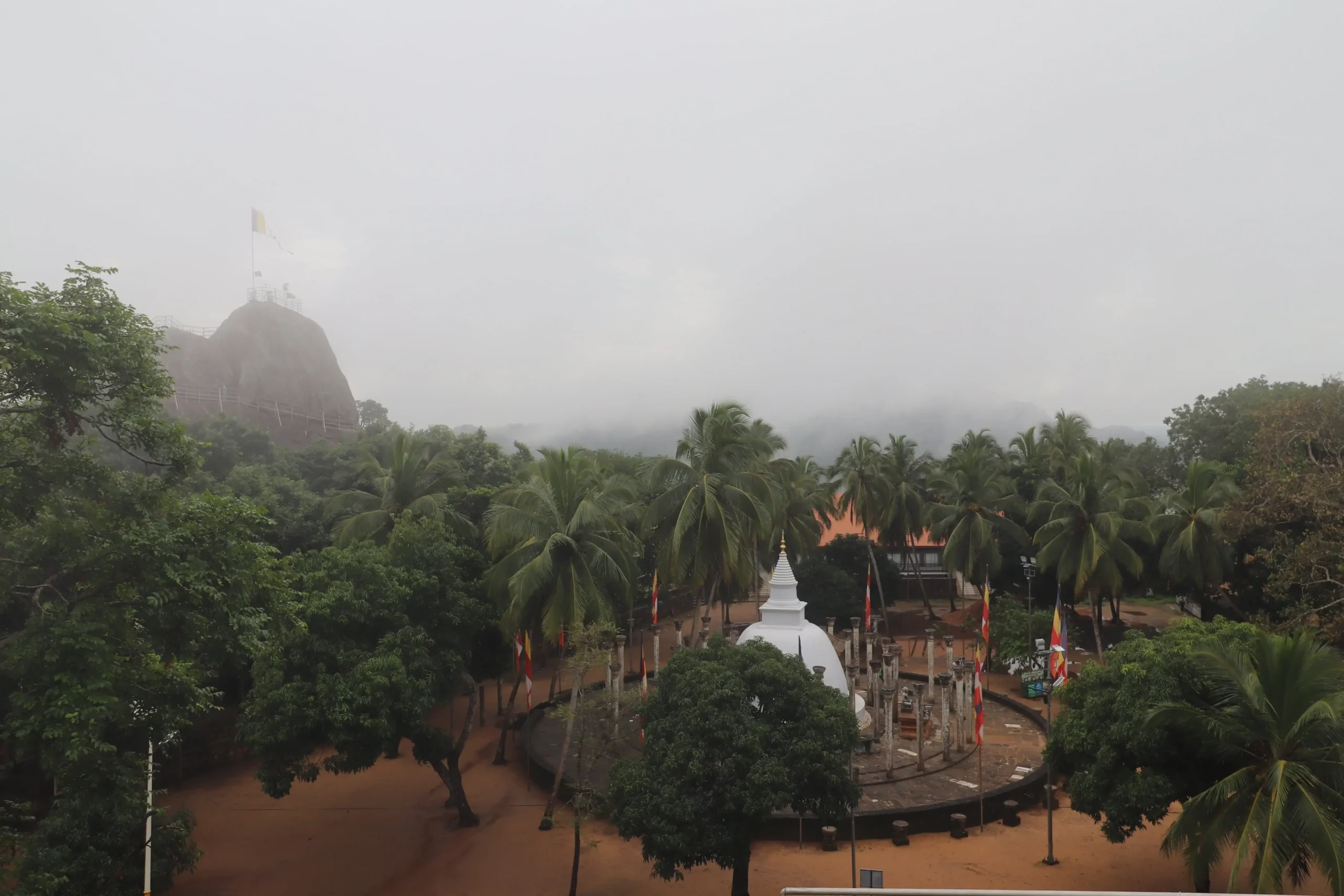 a misty viewpoint over Mihintale has a large rock with a flag overlooking palm trees surrounding a bright white bell-shaped dagoba, Sri Lanka