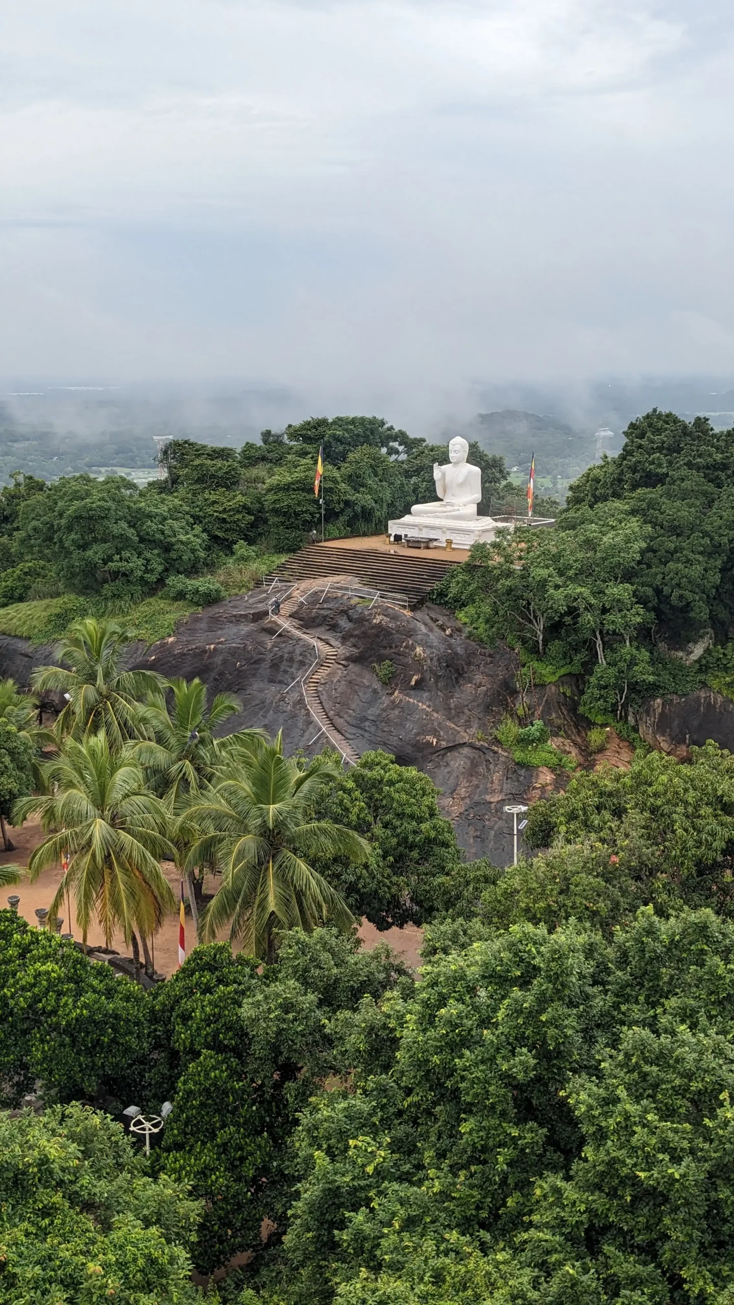 Mihintale a large, cross-legged, white statue of a Buddha is sat on a platform on top of a rock surrounded by rainforest and palm trees in Mihintale, Sri Lanka