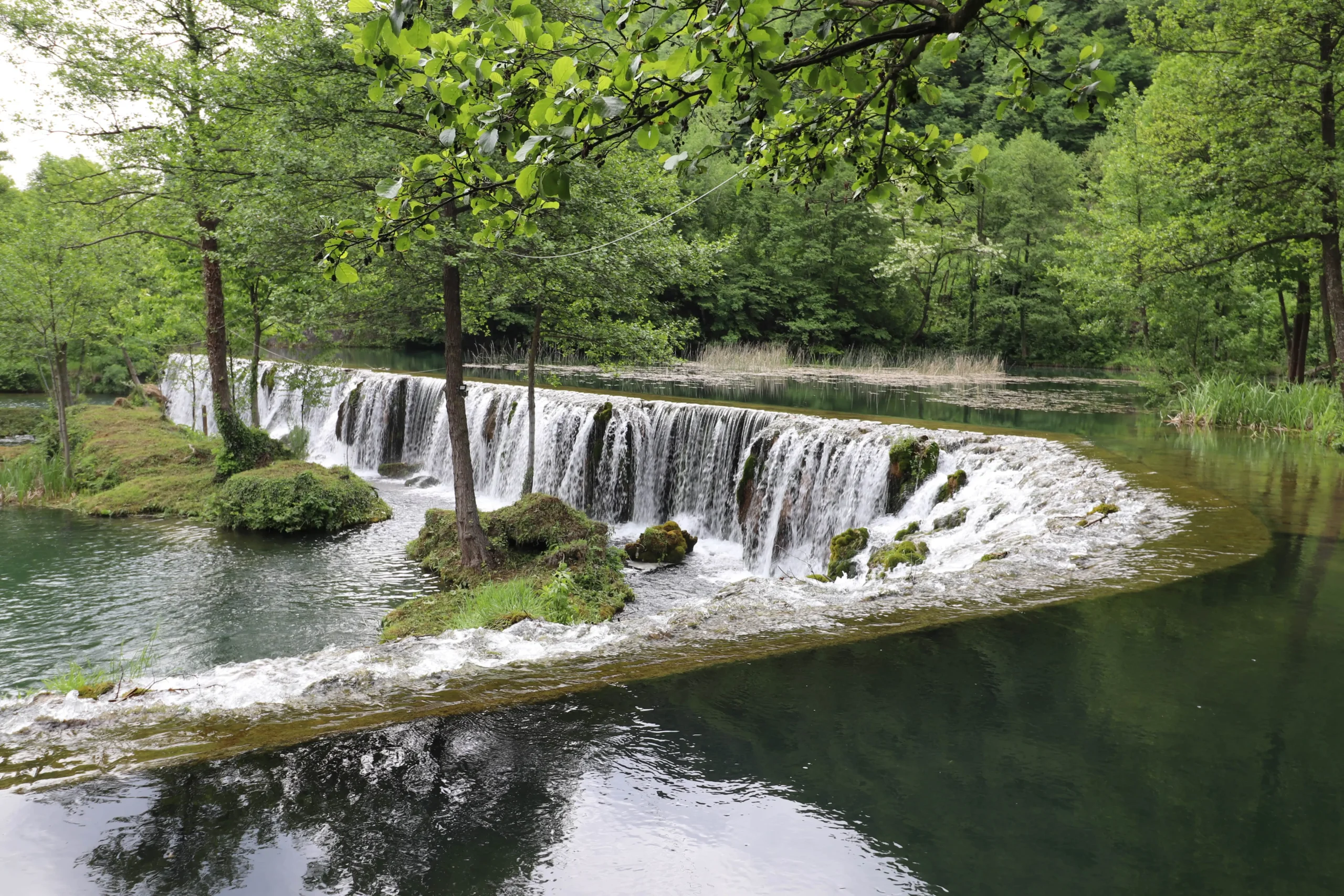 a short, wide and curved waterfall has small islands with trees growing at Konoba Slapovi in Jajce, Bosnia and Herzegovina