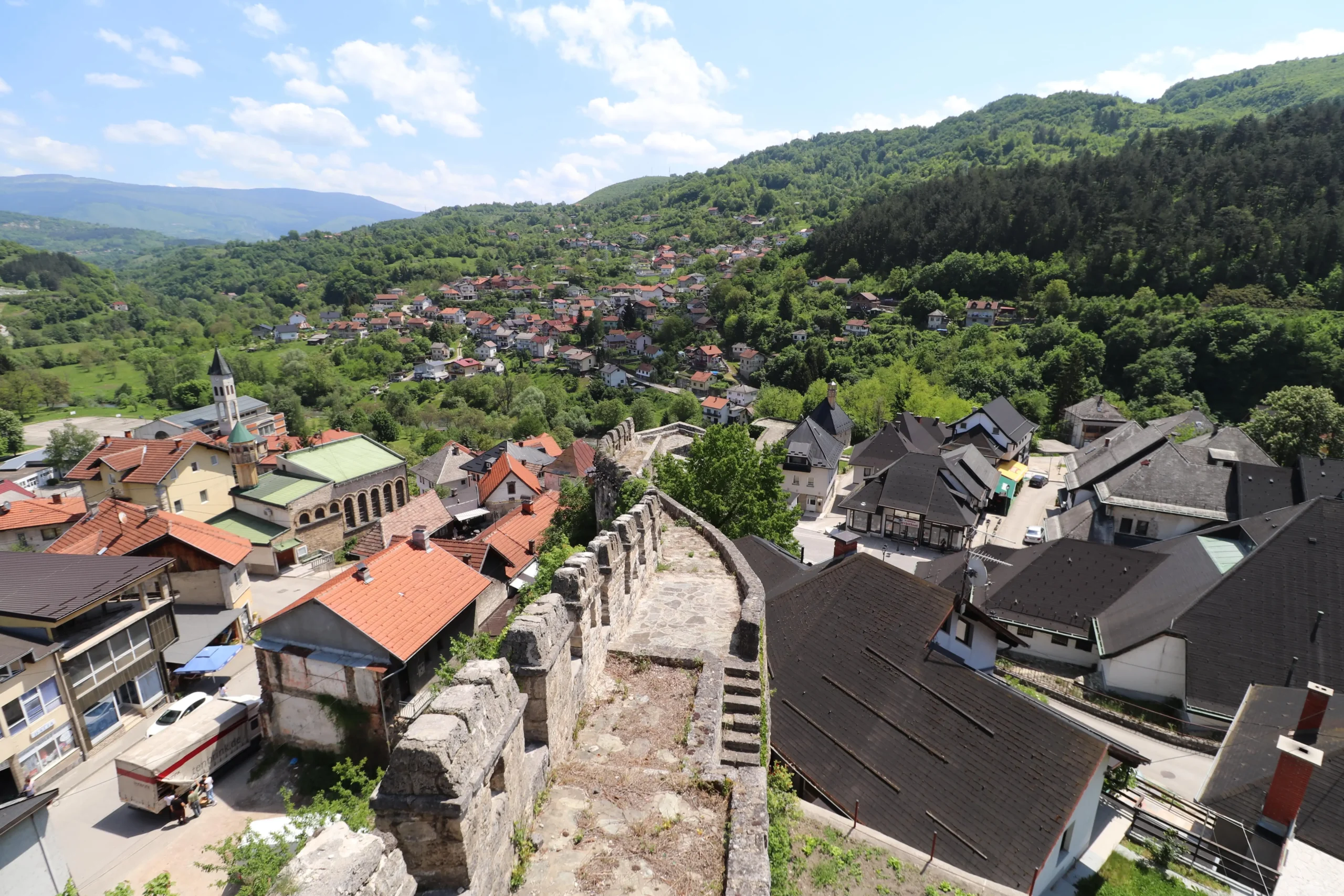 a view from Jajce Fortress over the town shows the fortress walls in the foreground, and rolling green hills dotted with orange roof-topped buildings - Bosnia and Herzegovina. Is Jajce worth visiting?