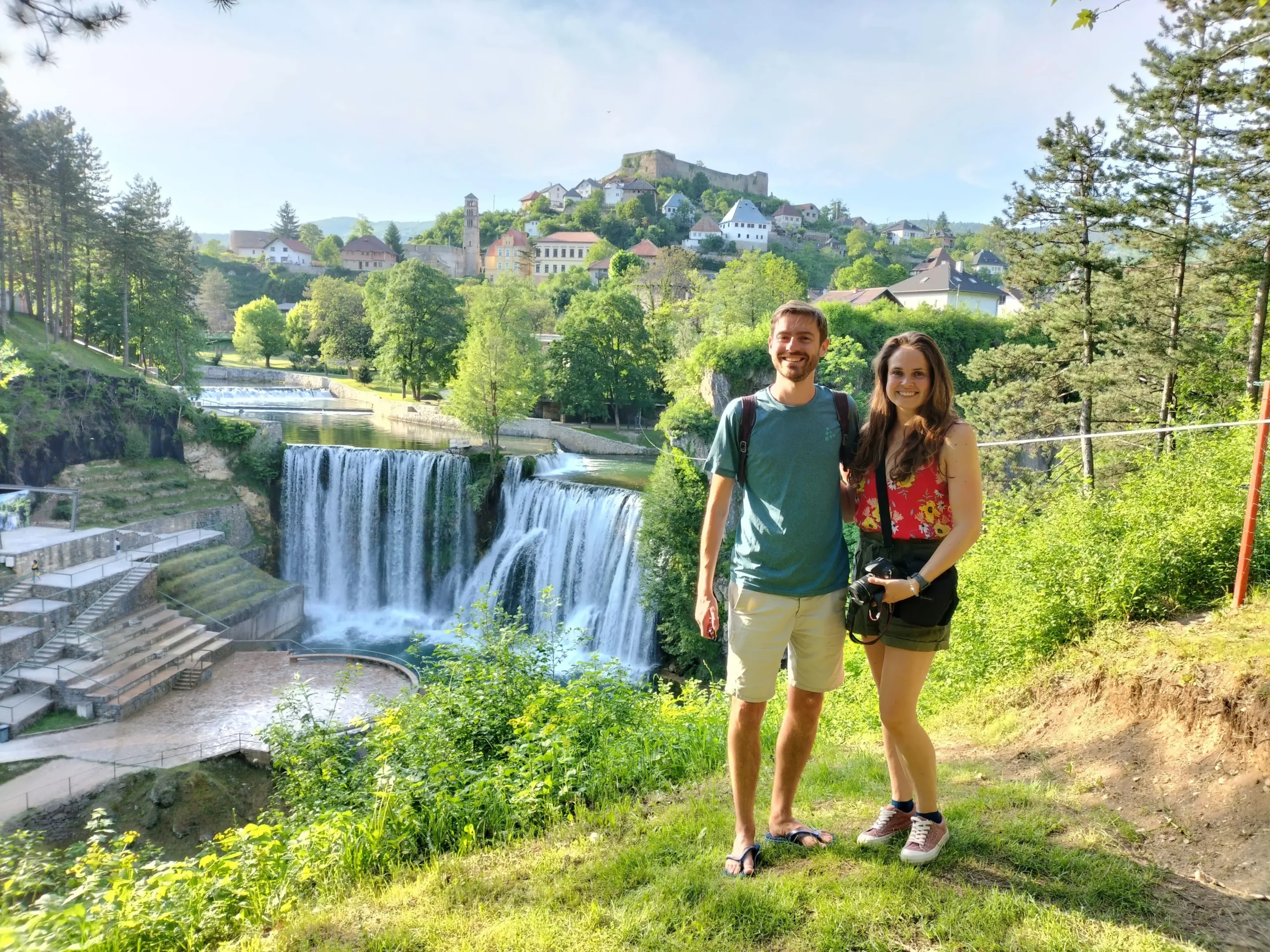 two people standing at a hidden viewpoint with an amazing view over the canyon with a huge waterfall on the other side, and medieval buildings of Jajce up the hillside in Bosnia and Herzegovina
