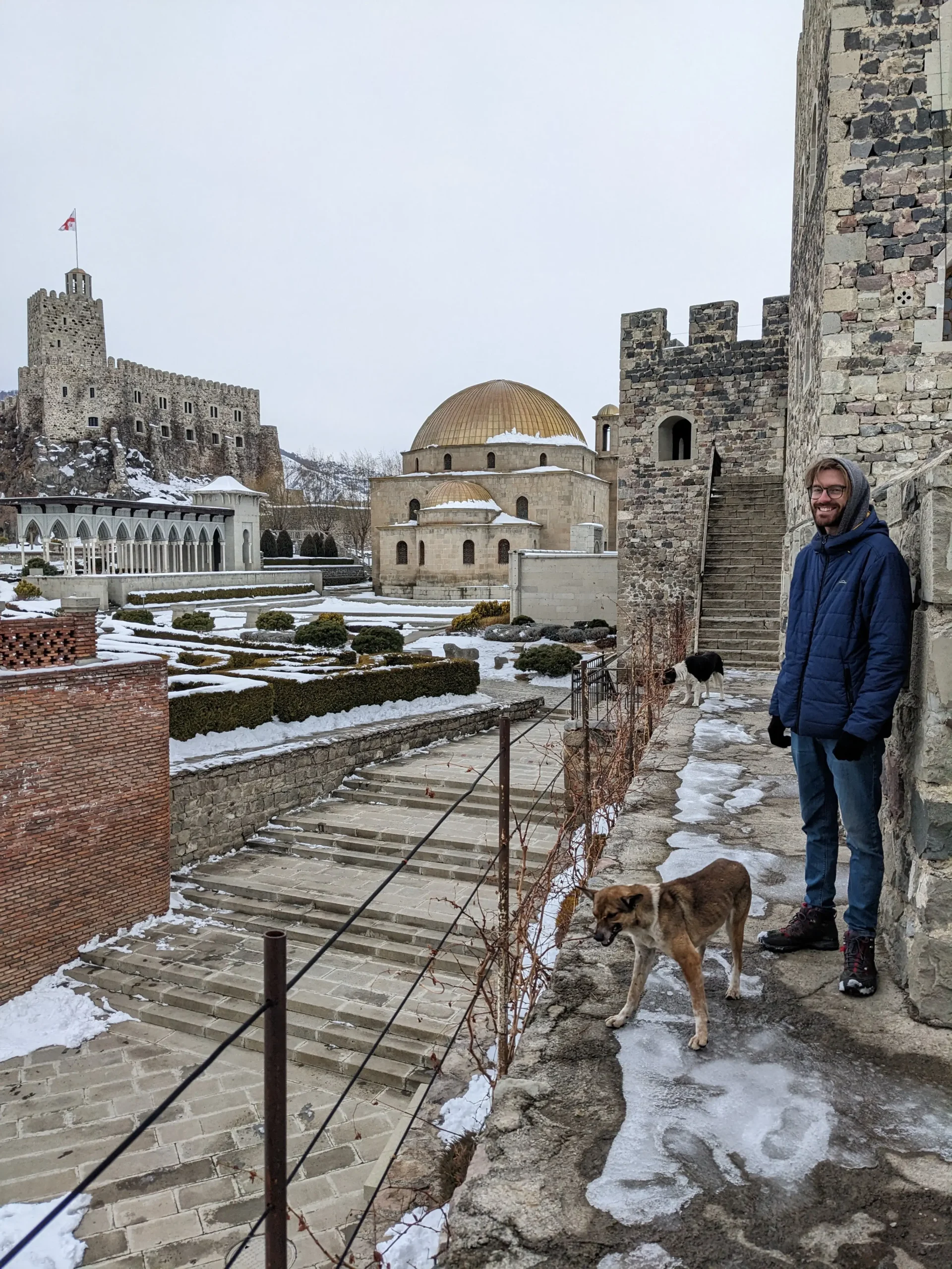 a man is standing on a wall of Rabati Castle with two stray dogs. There are brick fortress walls, a tower with a Georgia flag, and a golden domed mosque