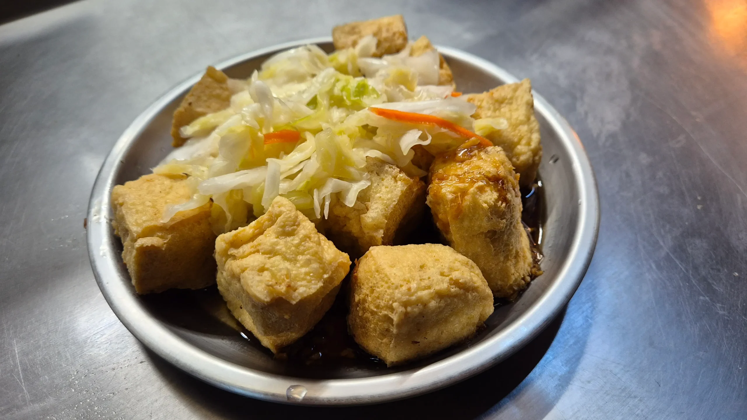 a metal bowl has deep-fried cubes of stinky tofu sitting in a soy sauce and topped with pickled cabbage at Nanjichang night market in Taipei, Taiwan