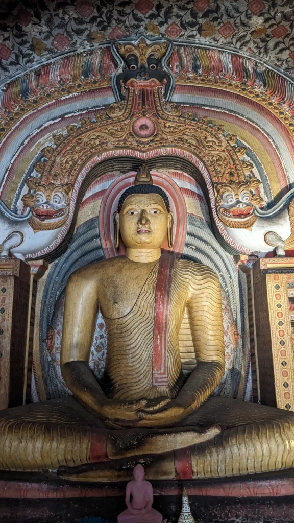 an ancient seated Buddha sculpture has intricate artwork on the wall behind depicting animals and flowers at Dambulla Cave Temple