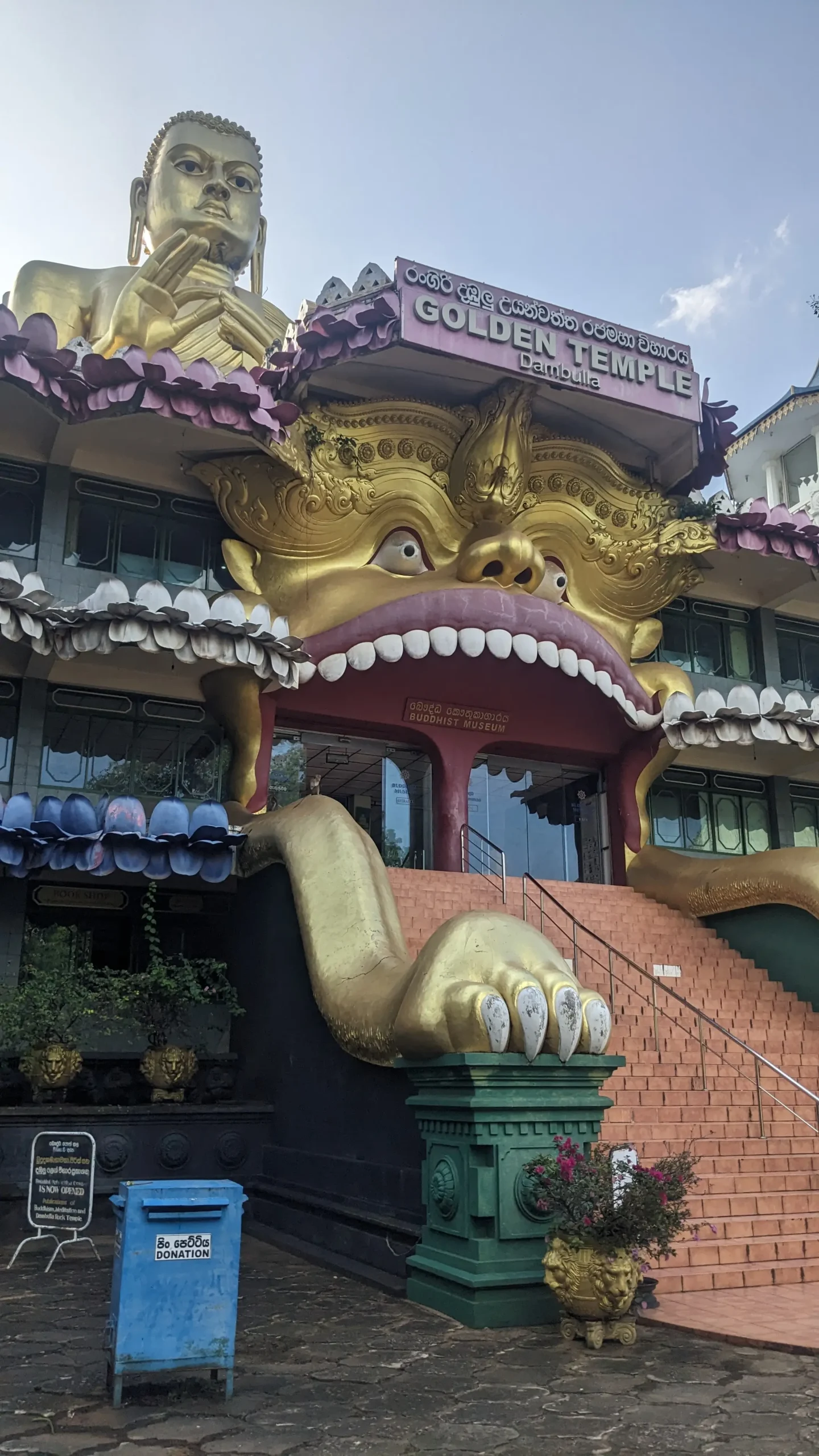 a Buddhist temple has a large gold seated Buddha statue on top and a a large face with an open mouth  around the entrance to the temple - Golden Temple, Dambulla