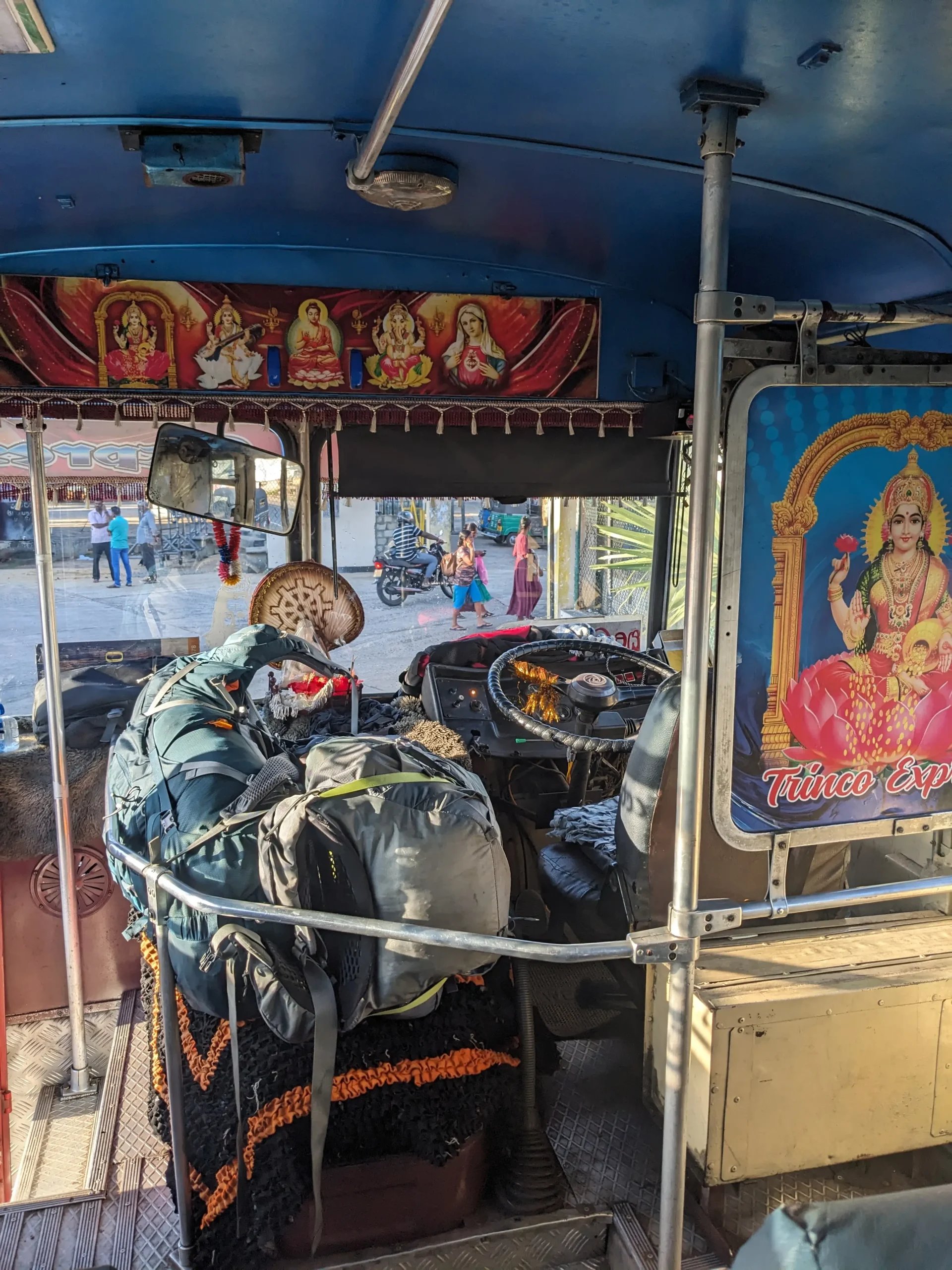 inside a Sri Lankan bus, the photo shows the driver's seat and two large backpacks strapped together next to it. There are posters on the walls of deities