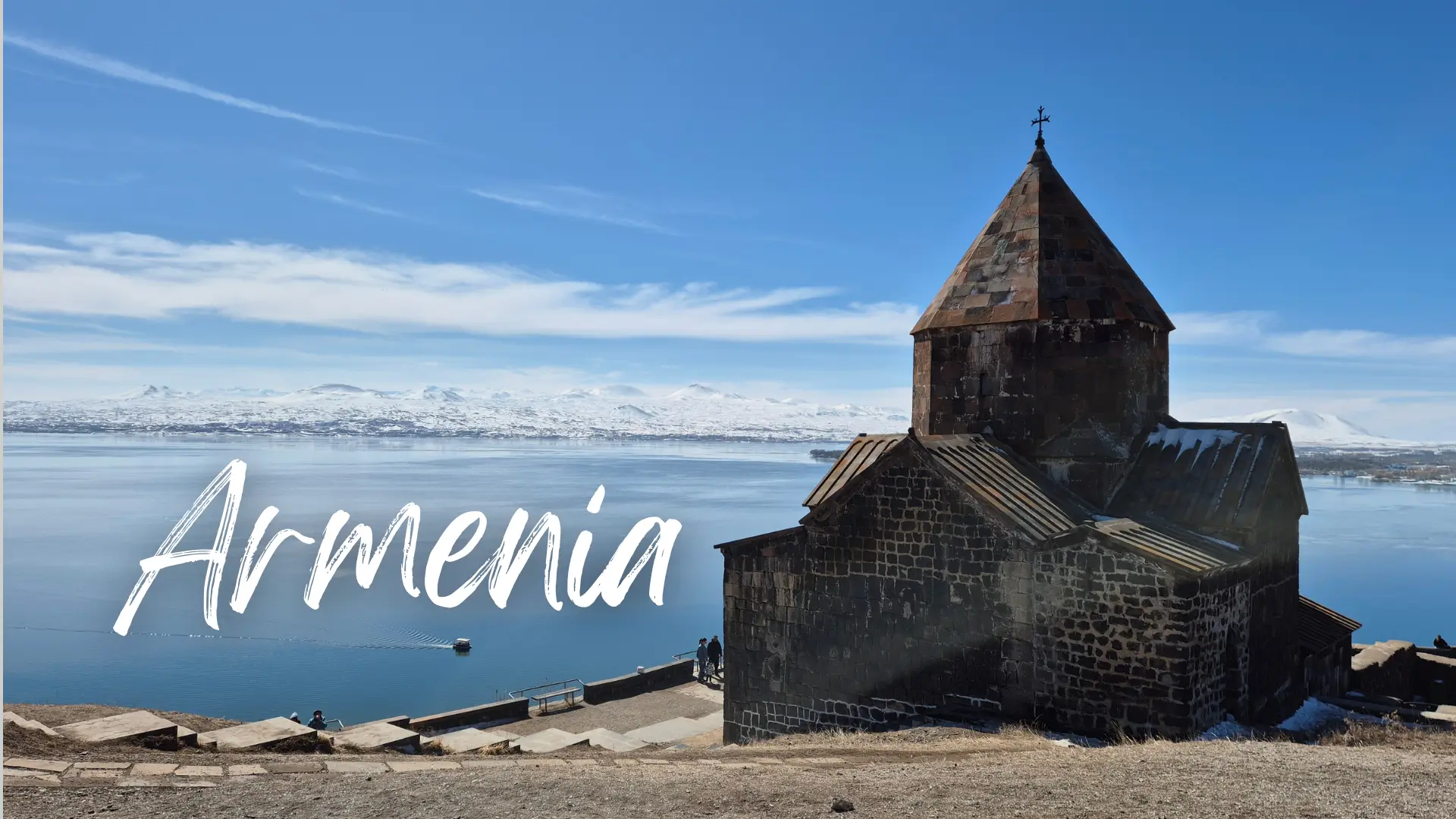 a historic brick monastery sits on a peninsula overlooking the large bright blue lake of Sevan with snow-capped mountains in the background. Armenia travel guide