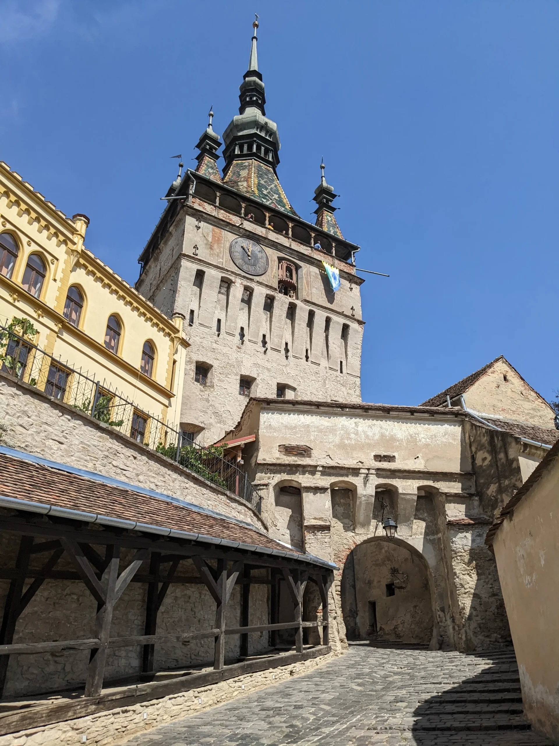 a cobbled street and covered walkway lead to a medieval archway and a 14th century clock tower with a patterned tiled roof and pointed spires. Things to do in Sighisoara