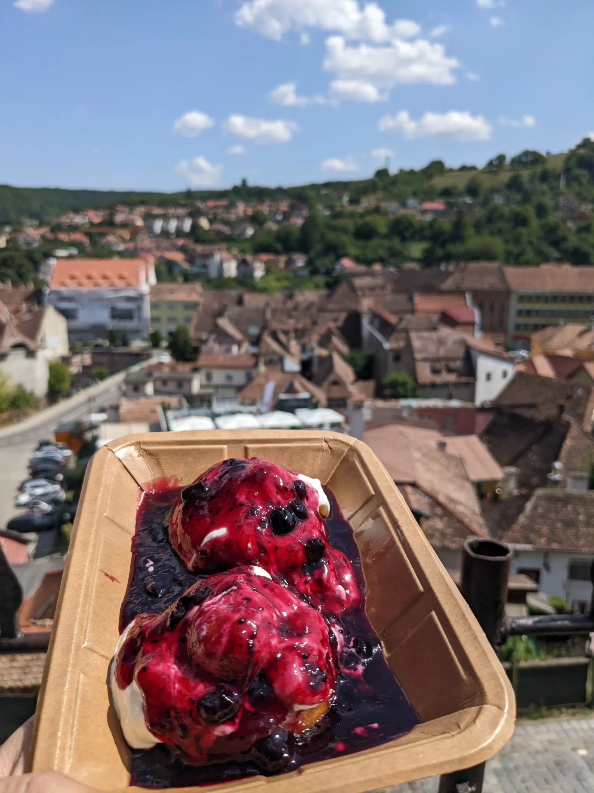 a container of two round cottage cheese doughnuts topped with berry jam and sour cream is held up with a view over Sighisoara town in the background